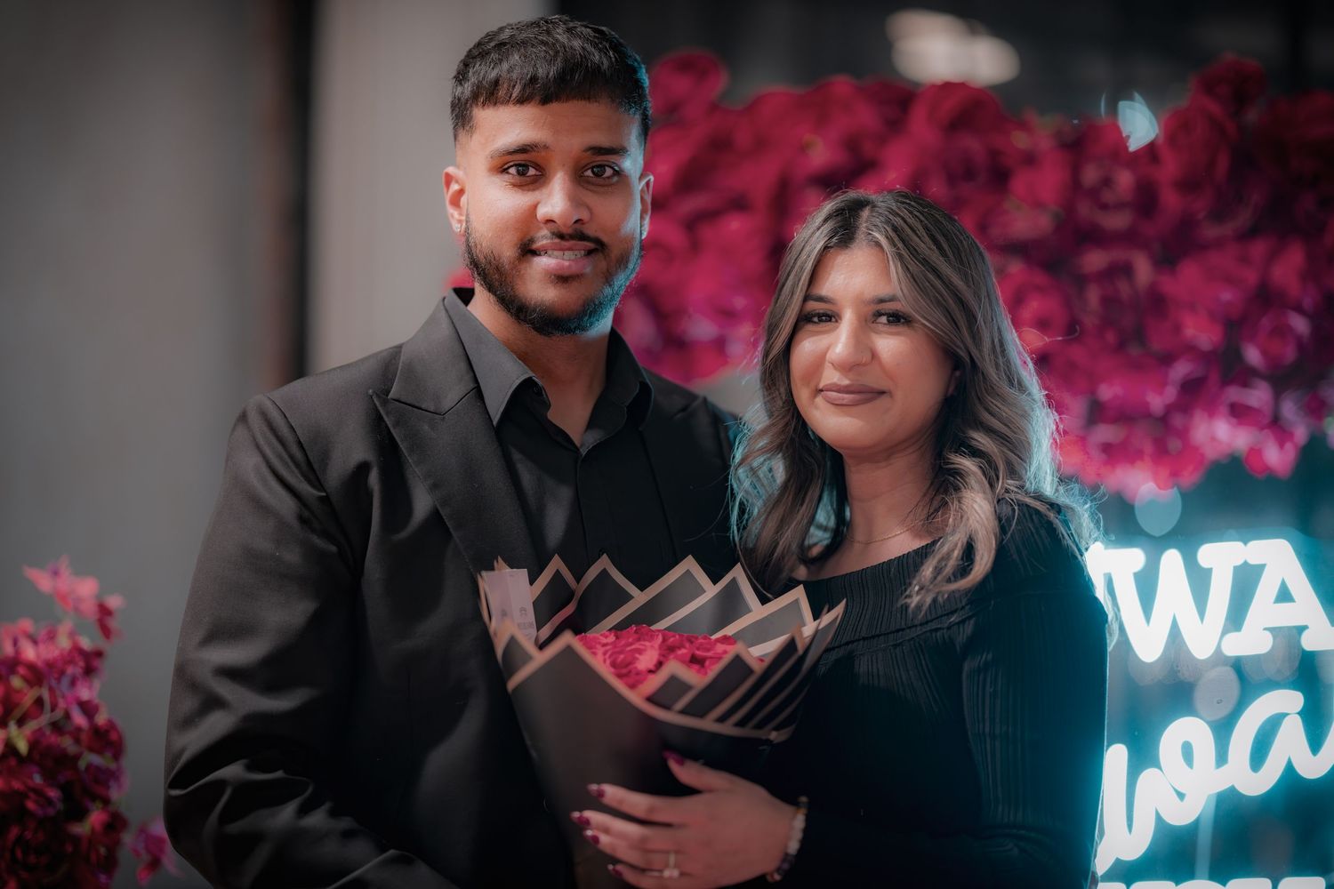 Romantic engagement scene with neon sign and red rose wall decorations.