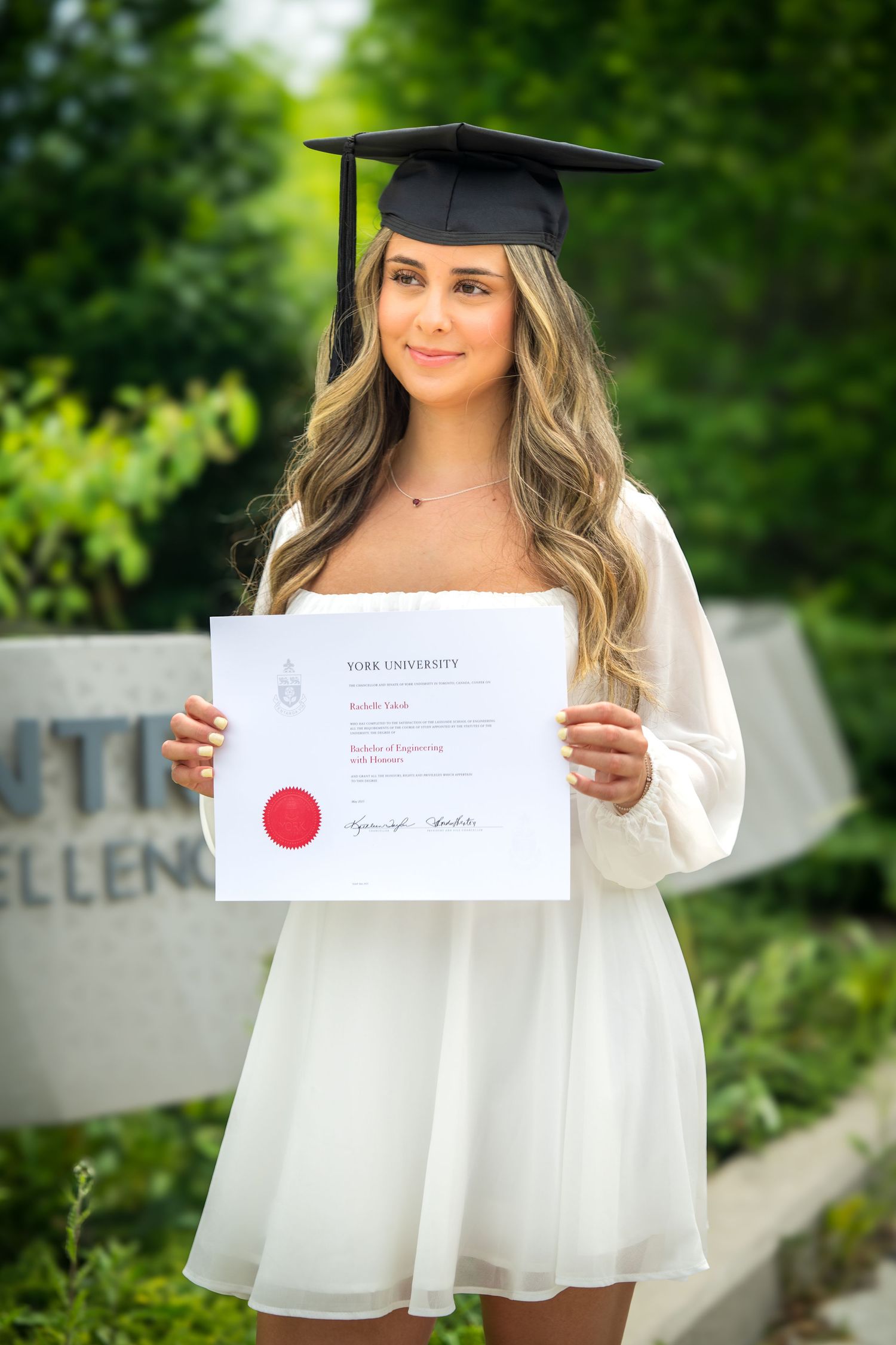 Graduate in white dress poses with diploma near university signage.