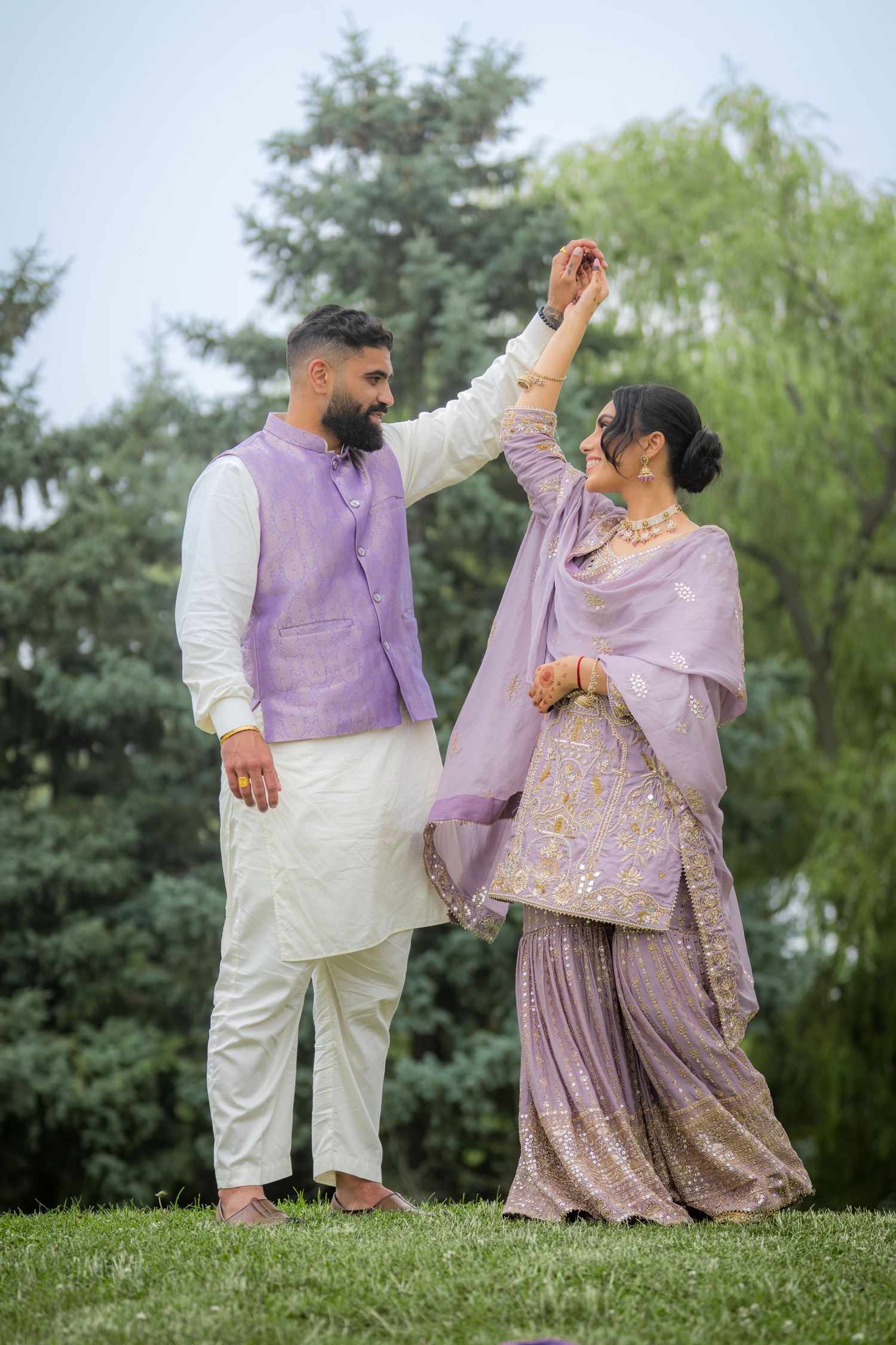 Couple in matching lavender Indian attire dancing outdoors, man twirling woman in embroidered lehenga among lush green trees.