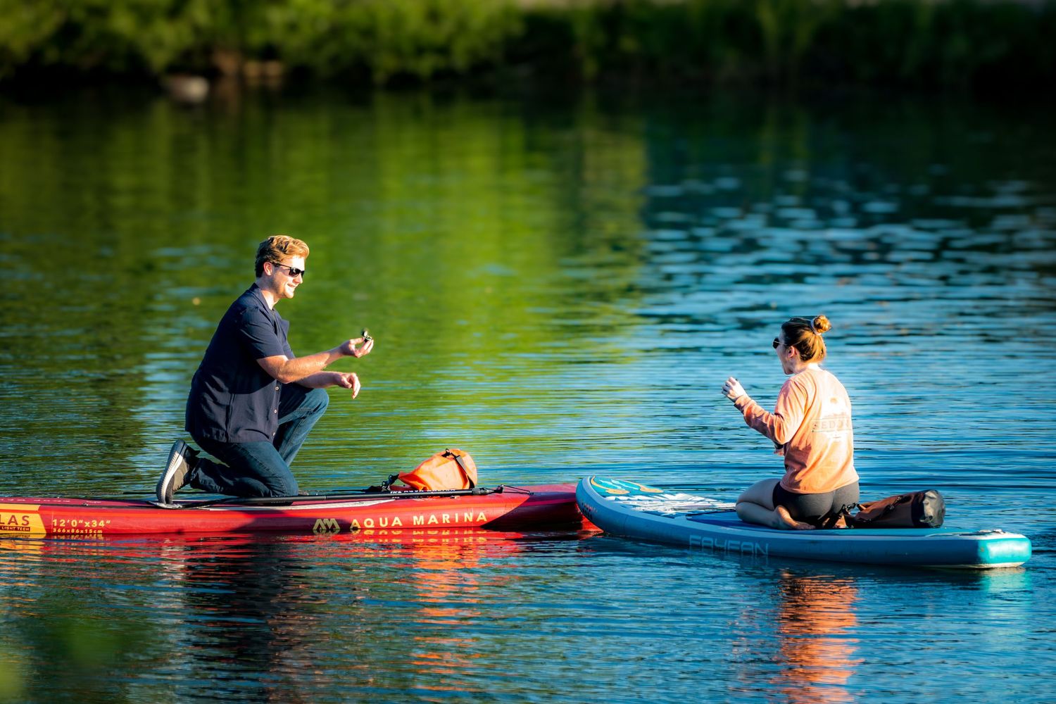 Two people kneeling on colorful paddleboards on a calm green lake, appearing to share a romantic moment.