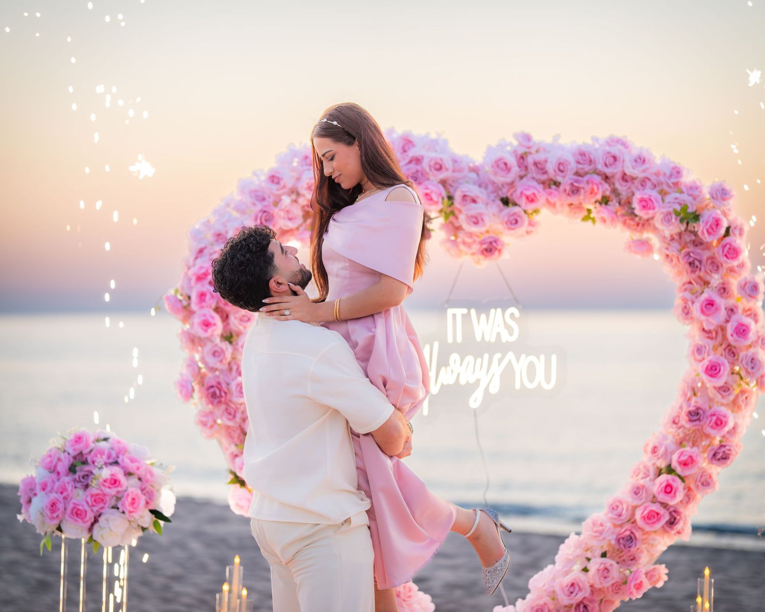 A couple embraces during a beach proposal under a pink floral heart arch at dusk.