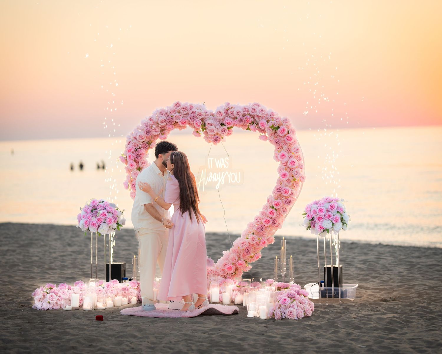 A romantic beach scene with a heart-shaped flower arch and glowing candles at sunset.