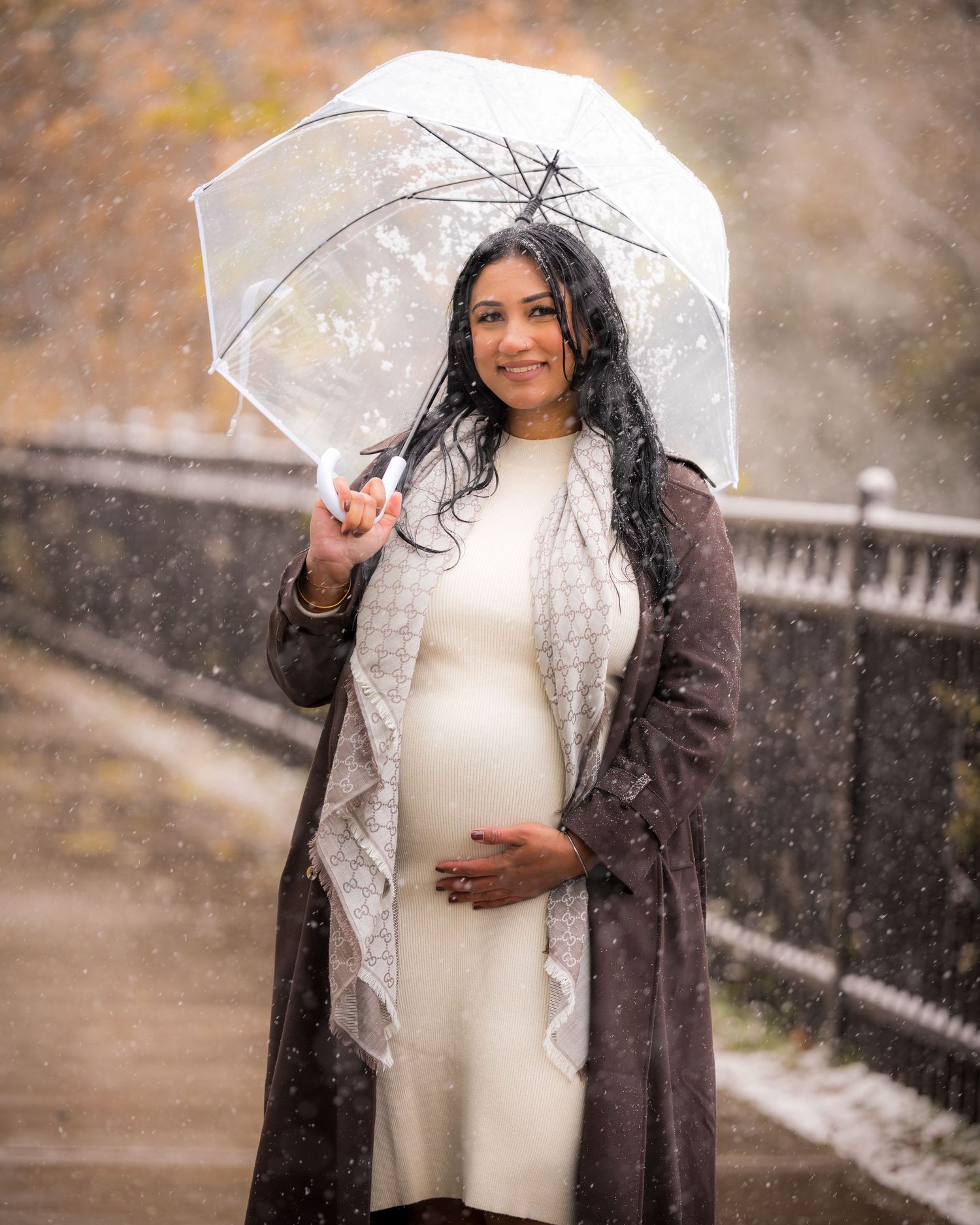 A pregnant person in winter attire poses with a clear umbrella during snowfall.