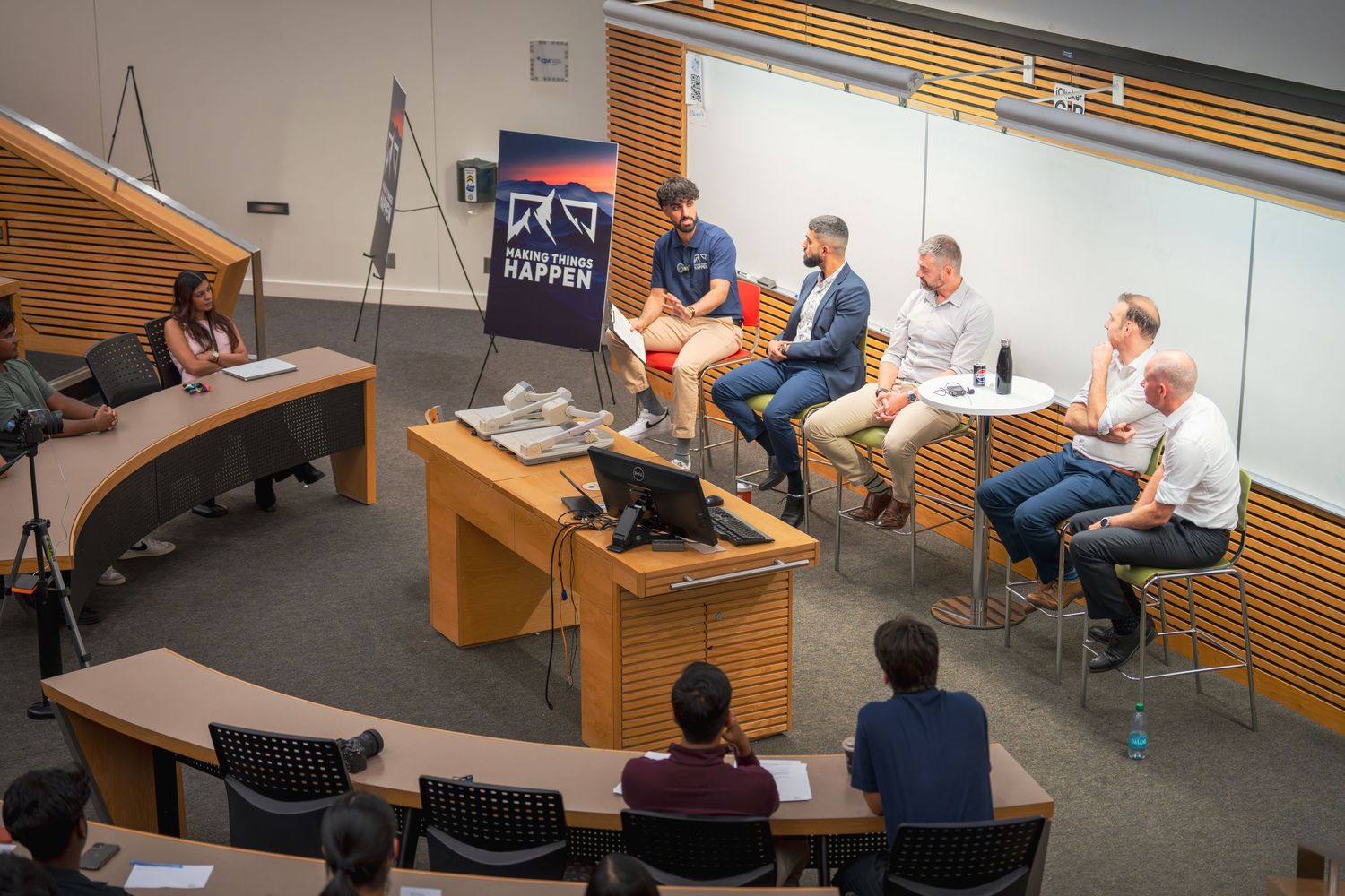 Wide shot of a panel discussion in a tiered classroom with audience members.
