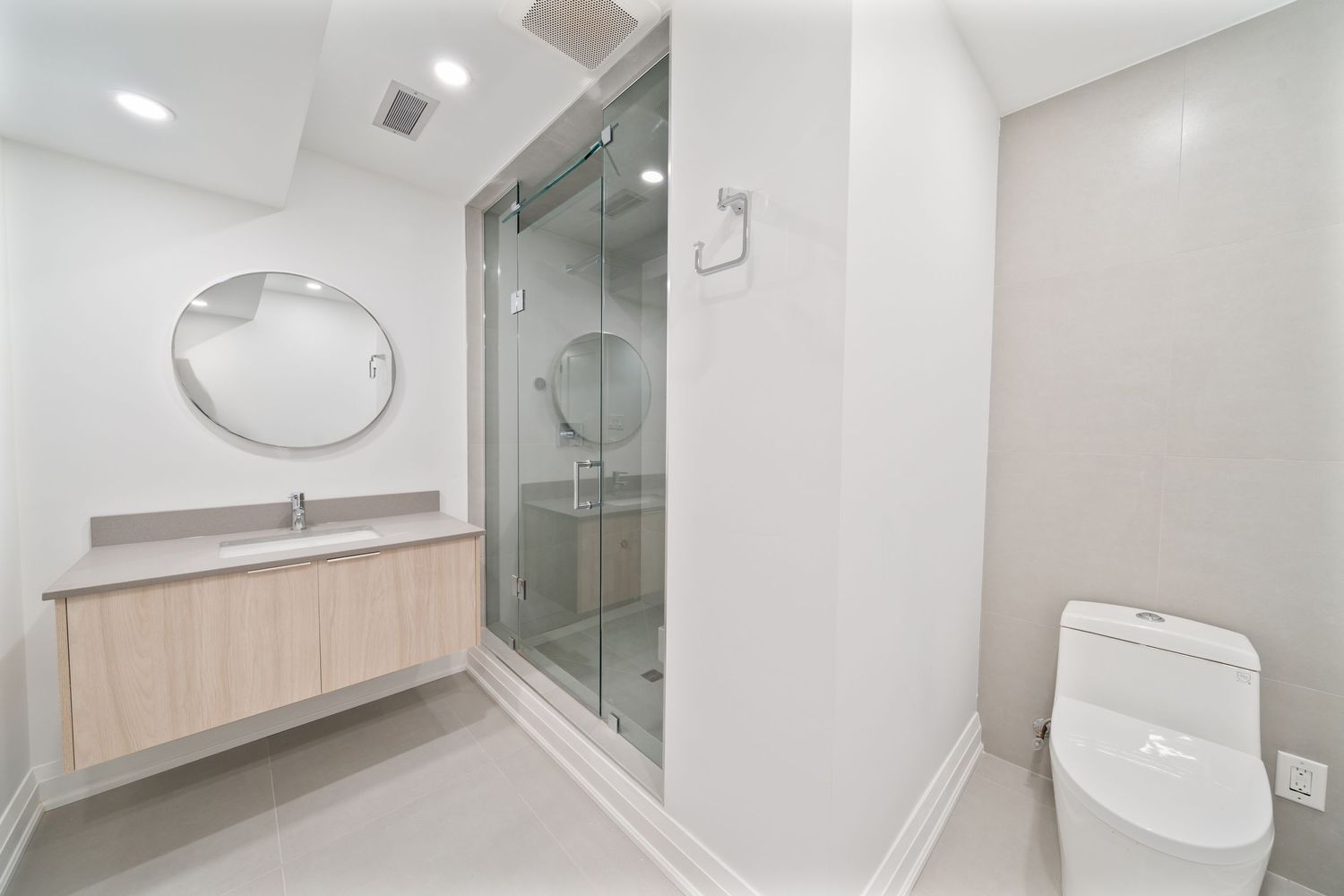 Modern bathroom interior with glass shower enclosure, light wood vanity, oval mirror and gray tile flooring.