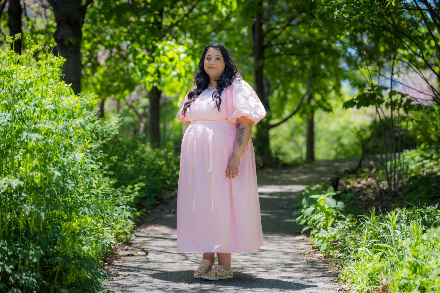 Woman in flowing pink maxi dress standing on a stone path in a sunny garden.