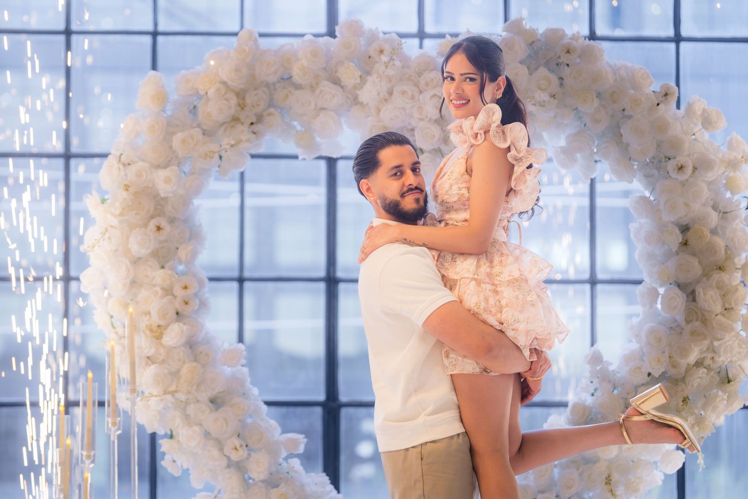 Groom lifts smiling bride in front of large white floral heart arch backdrop.