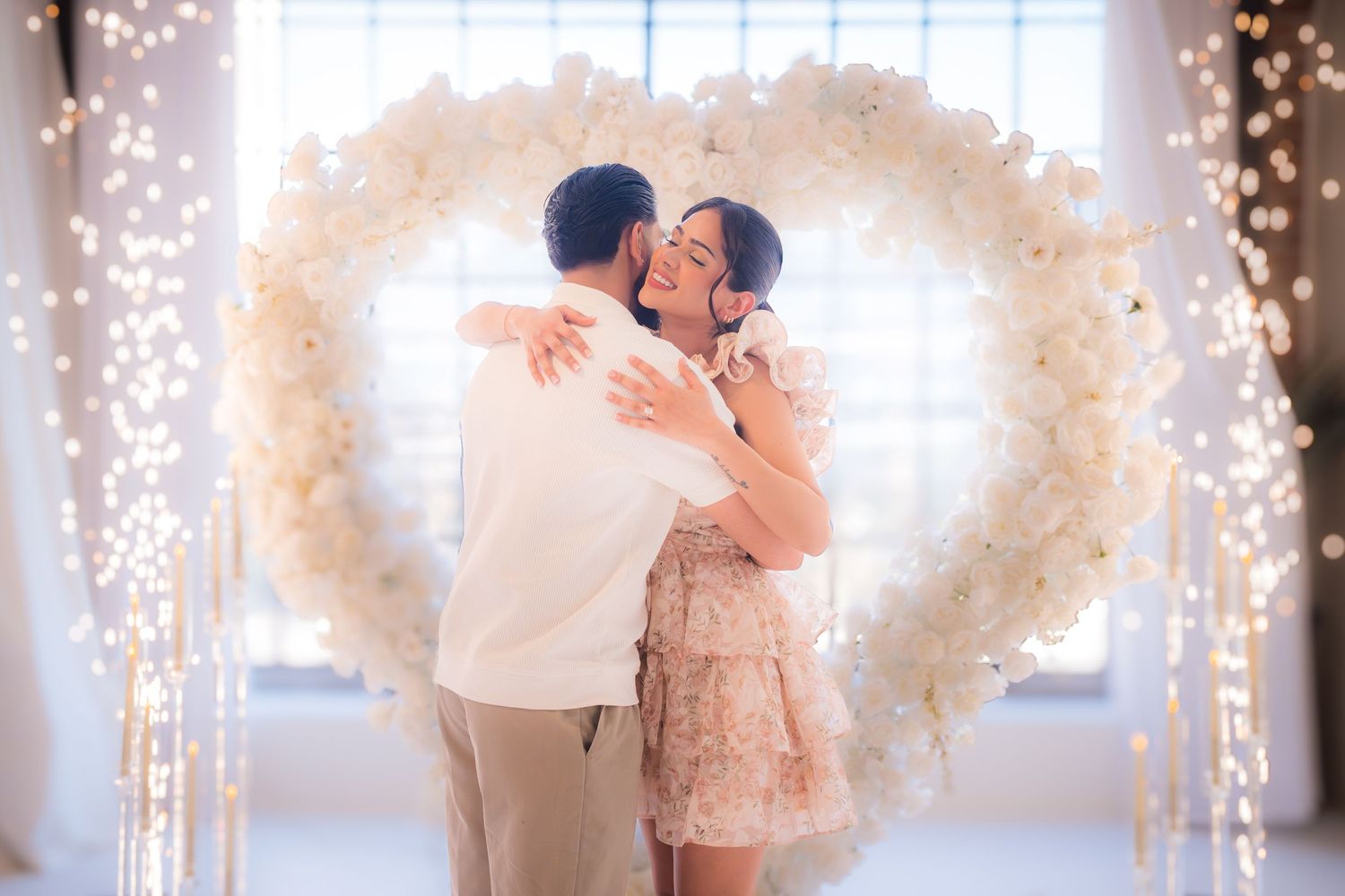 Happy couple embracing and kissing beneath glowing white floral heart arch backdrop.