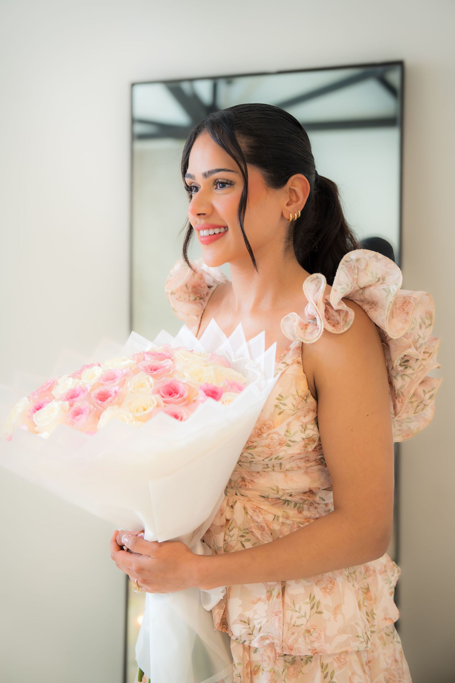 Smiling woman in floral dress holding pink and white rose bouquet in front of mirror.
