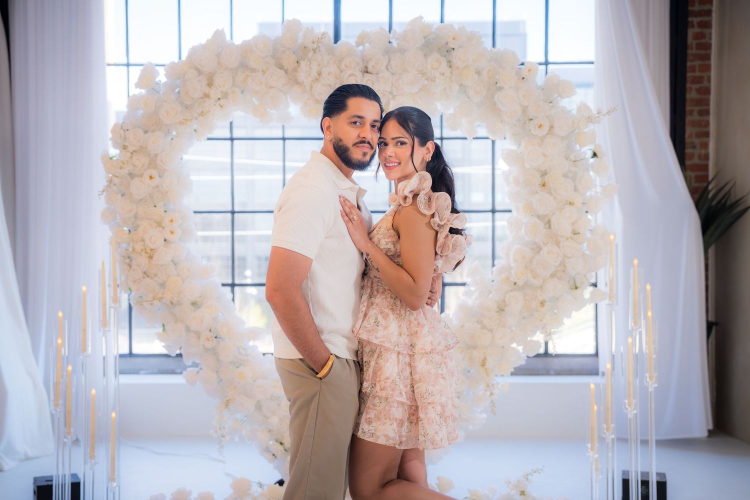 Smiling engaged couple posing in front of a large white floral heart arch with candles.