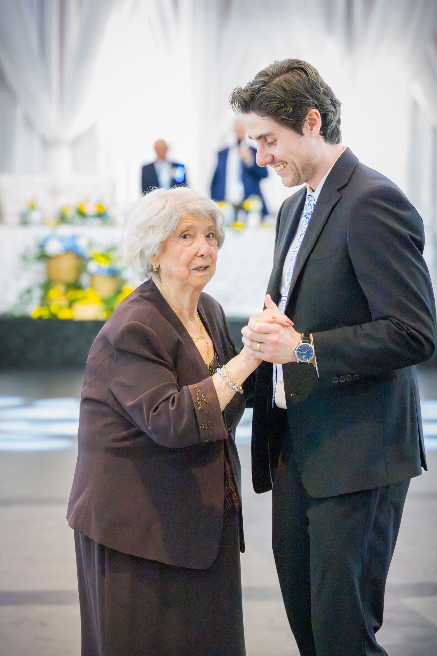 Elderly woman dancing with young man in dark suit at formal celebration event.