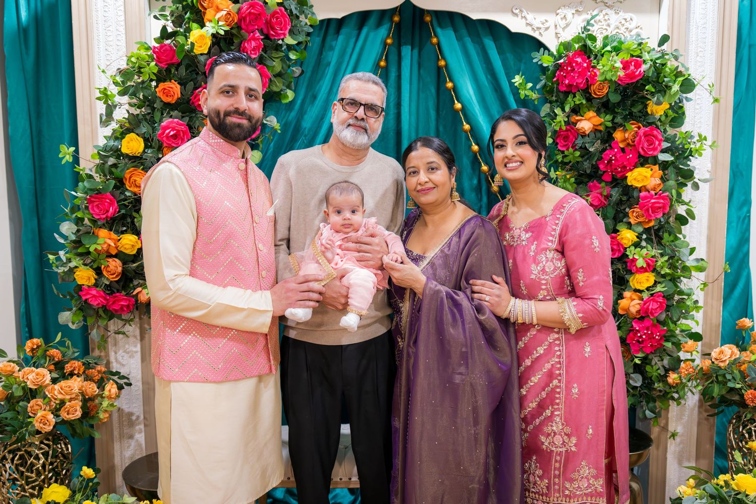 Five adults including grandparents posing with newborn baby at colorful floral decorated Indian celebration backdrop.