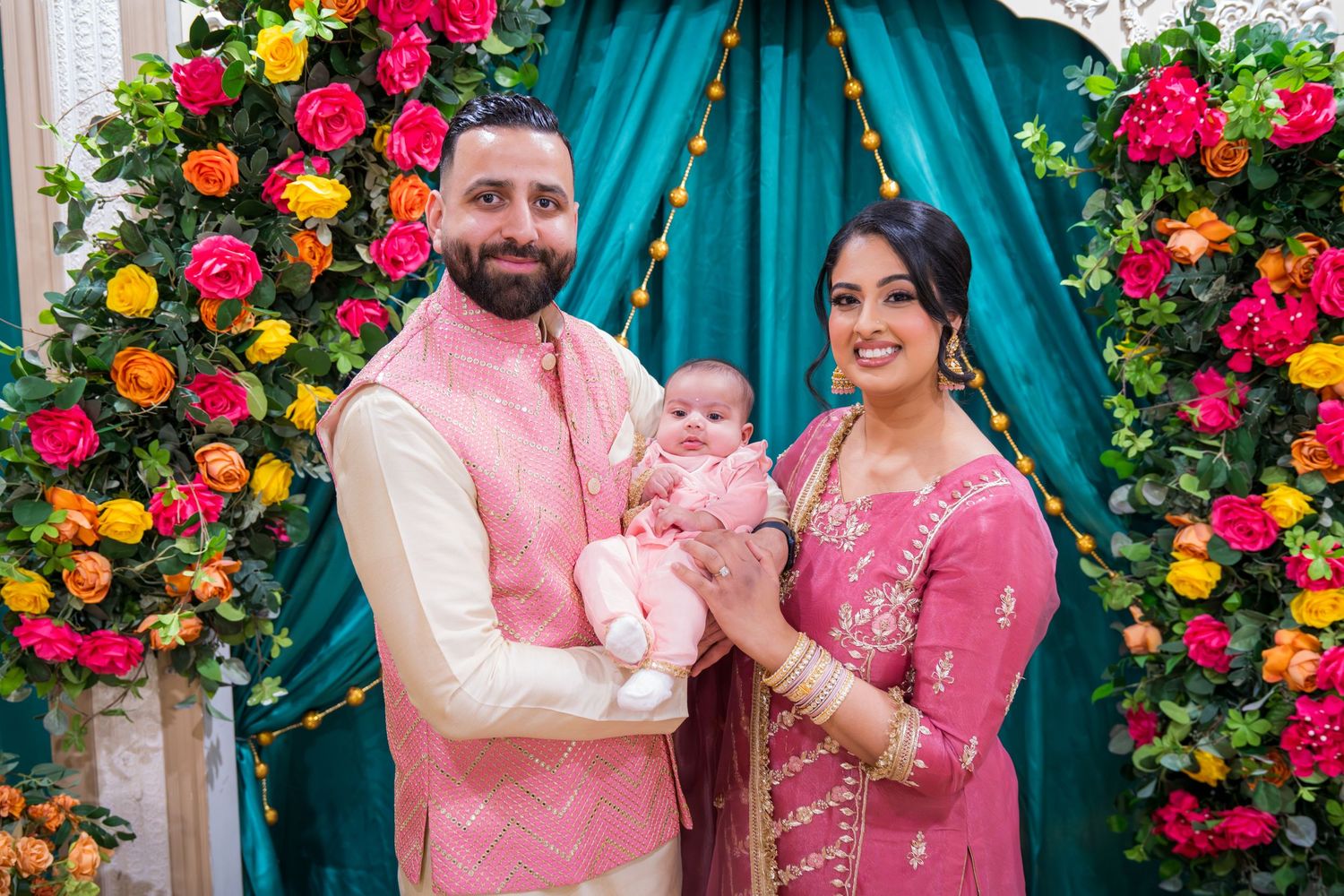 South Asian family in pink traditional attire holding newborn baby at floral decorated ceremony.