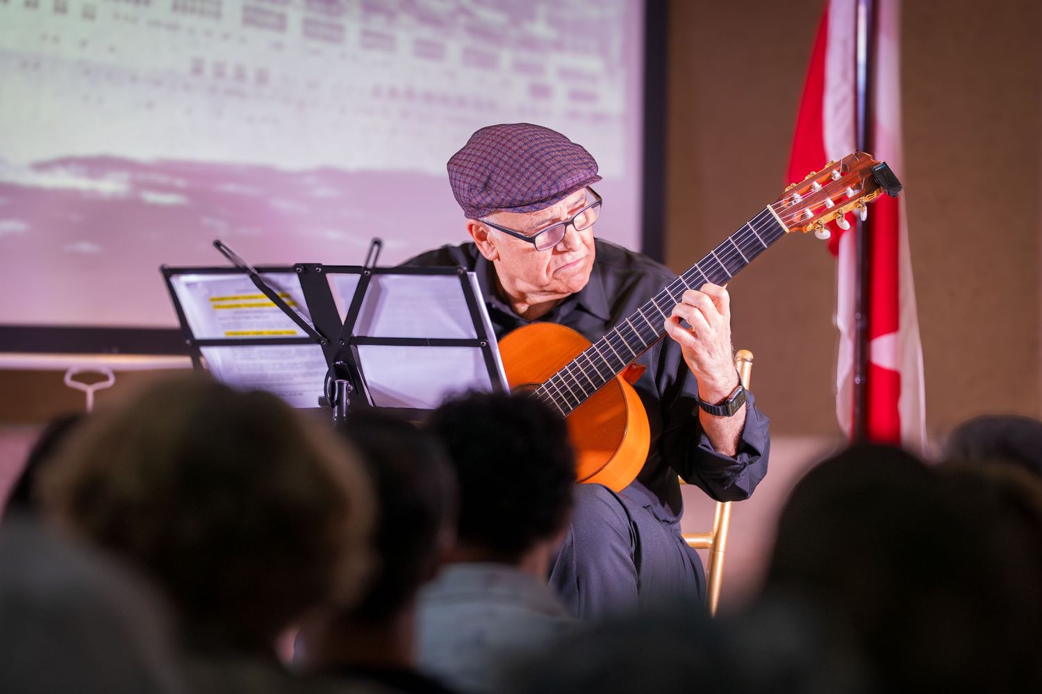 Senior male guitarist in cap playing classical guitar before an audience at an indoor concert.