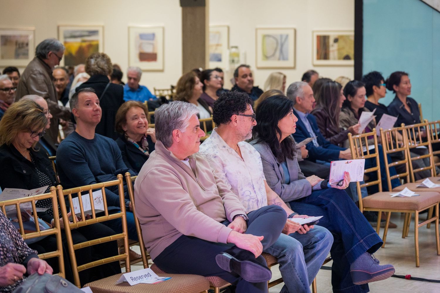 Audience members seated in wooden chairs attentively watching a live performance in an art gallery.