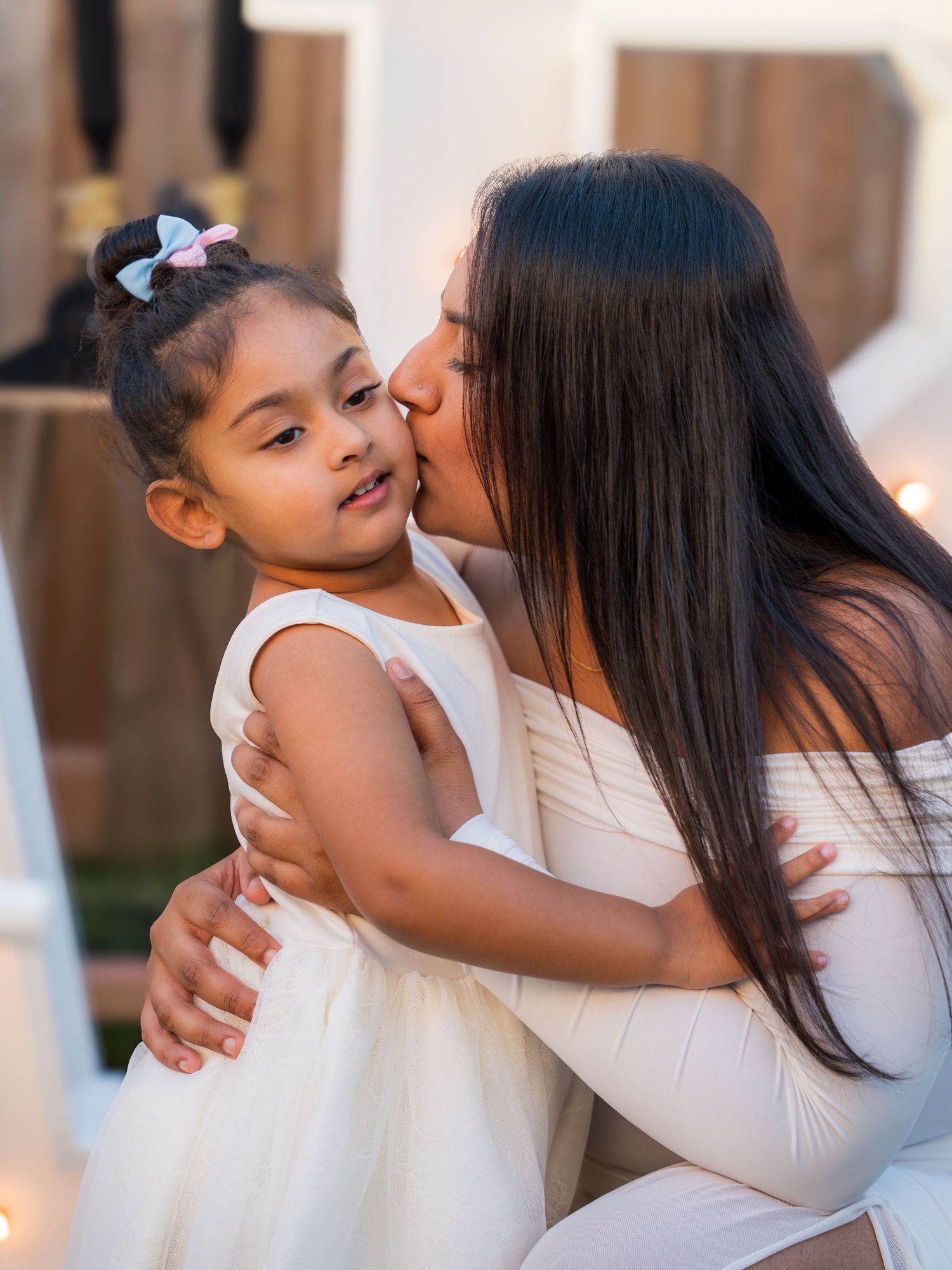 A mother in white kisses her young daughter on the cheek at a festive outdoor event.