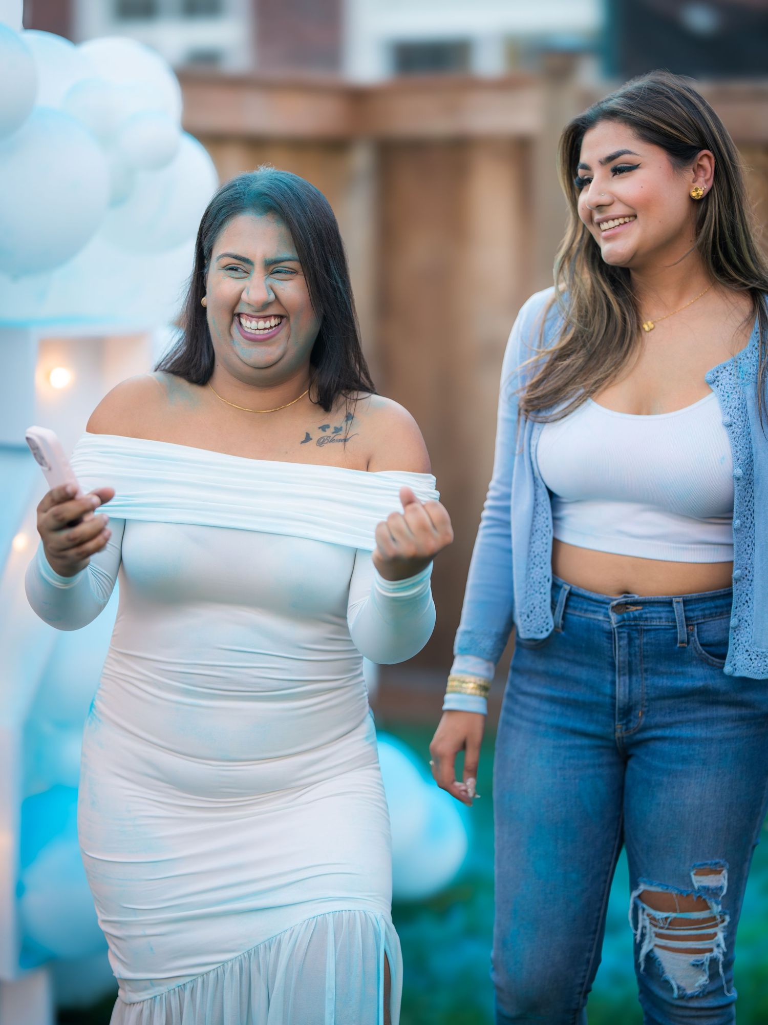 Two women laugh joyfully outdoors near white balloon decorations at a gender reveal celebration.