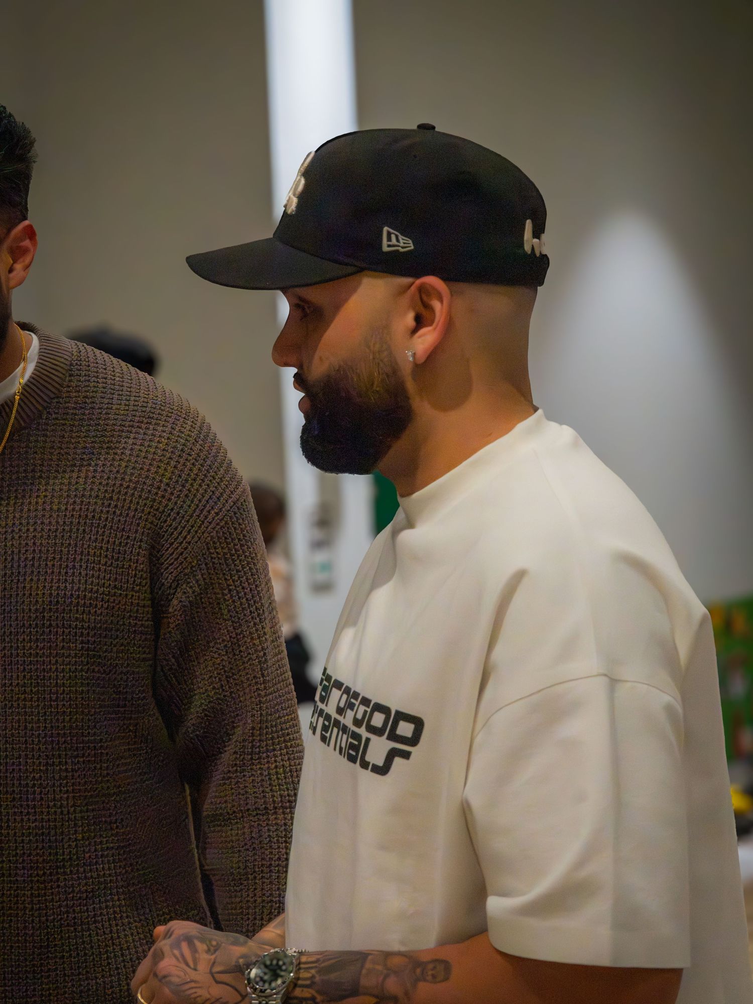 A bearded man in a white Fear of God Essentials tee and black cap stands in profile at an event.
