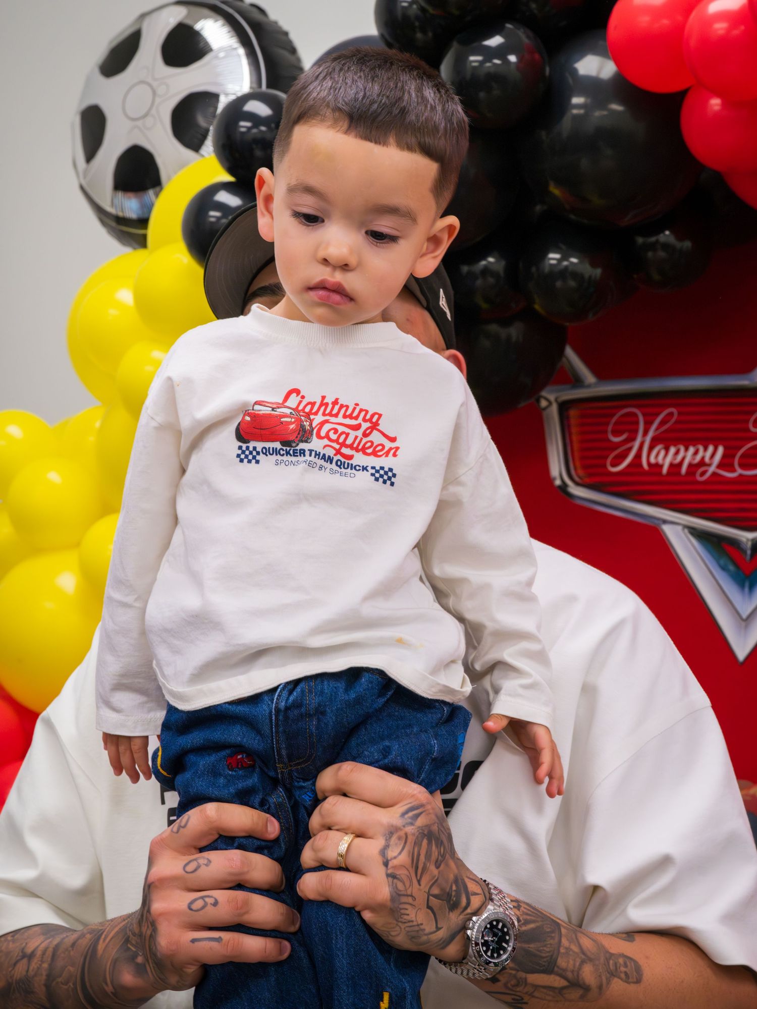 Toddler boy in Lightning McQueen shirt held up by tattooed hands at birthday party.