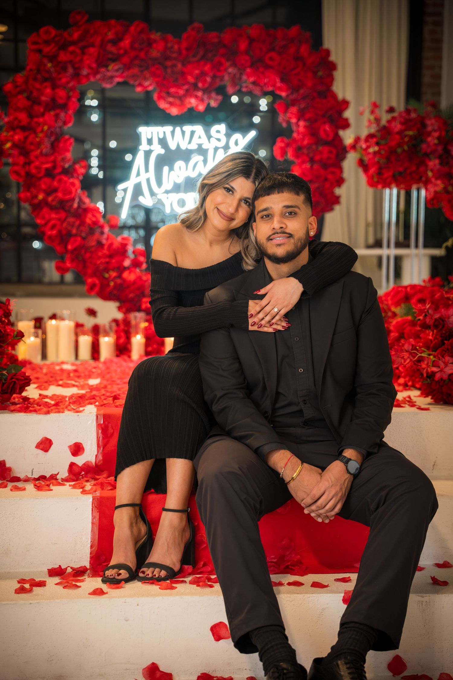 Elegant couple shares intimate moment surrounded by red rose wall and neon signage.