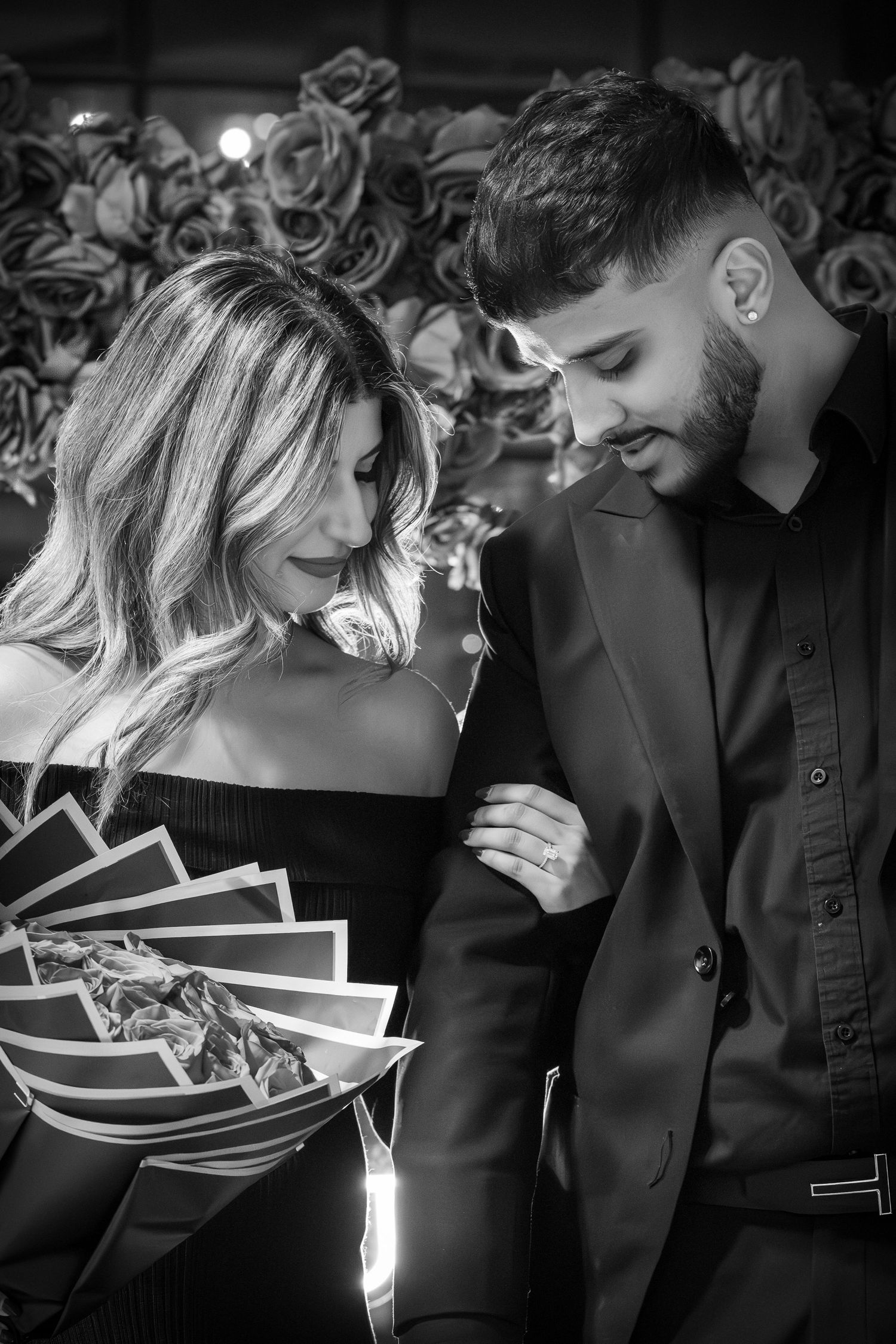 A romantic black and white portrait of a couple in formal attire sharing an intimate moment against a floral backdrop.
