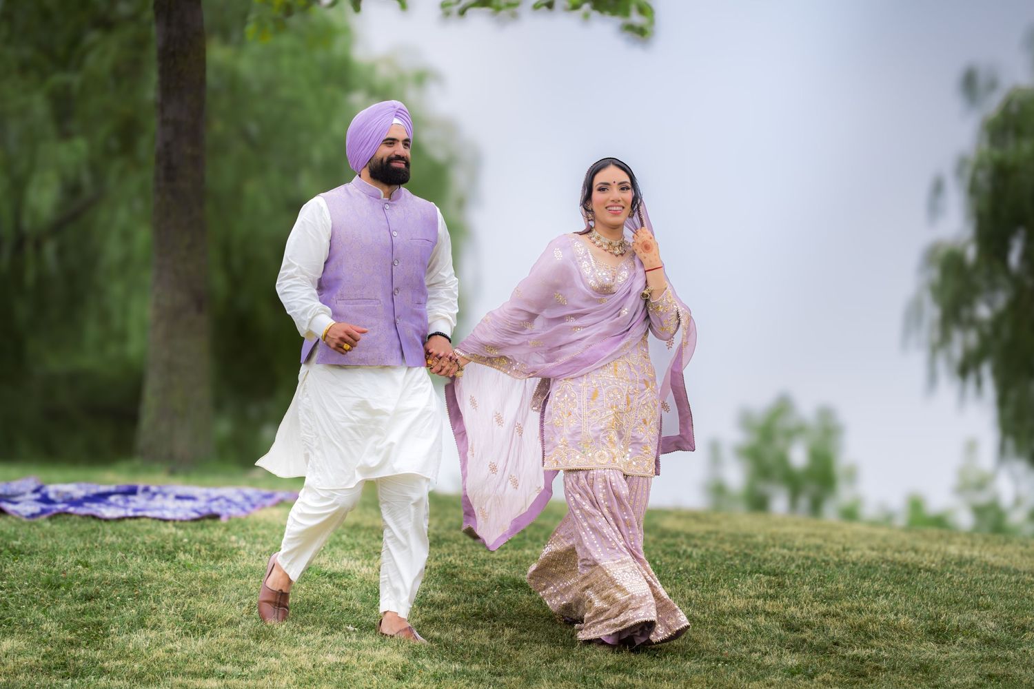 A couple in coordinated purple and white traditional clothing poses on a hillside during sunset.