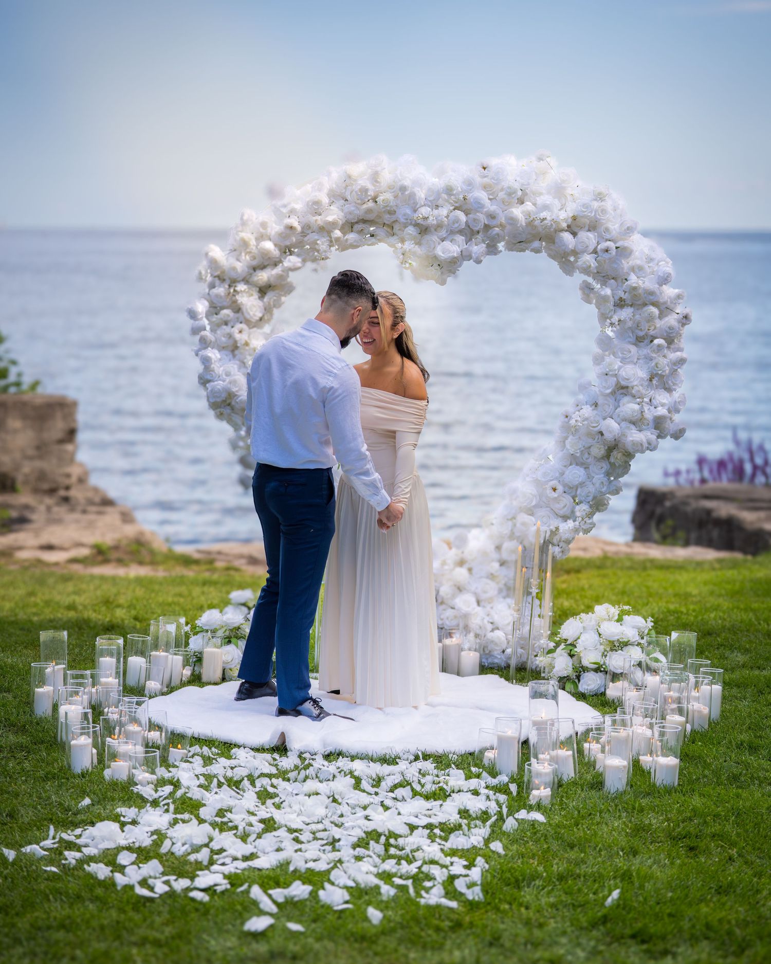 Intimate wedding ceremony by the ocean with white floral arch and scattered rose petals creating a romantic atmosphere.