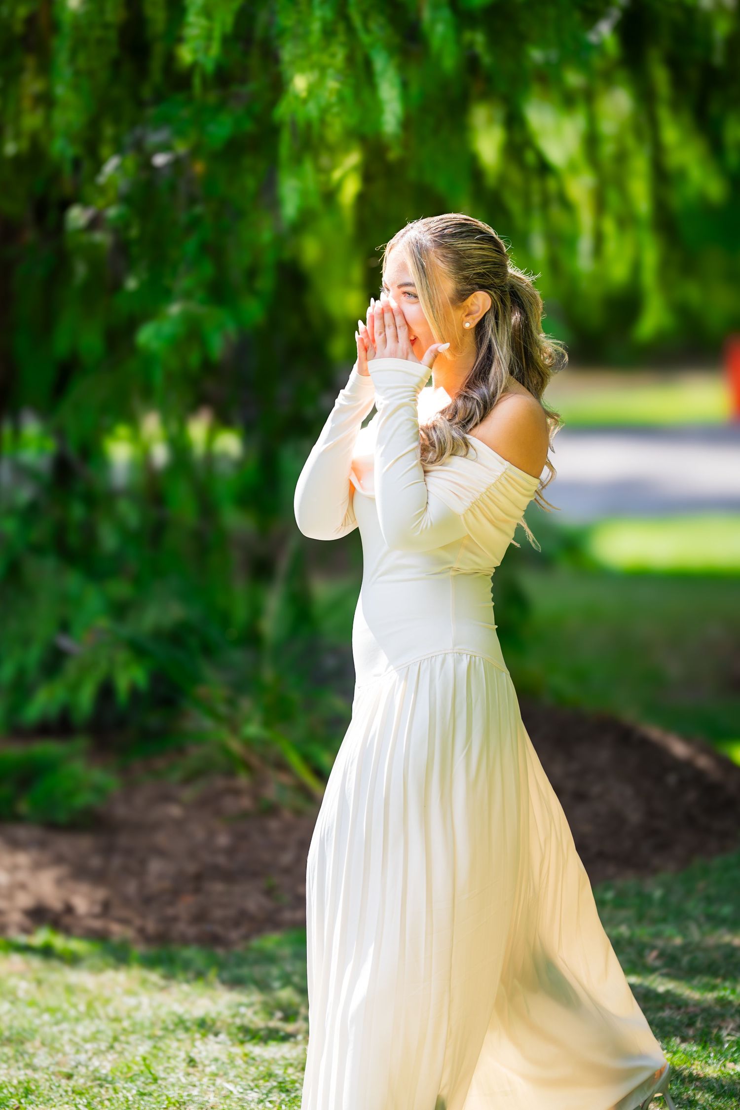 A person in an elegant white dress poses outdoors while covering their face playfully.