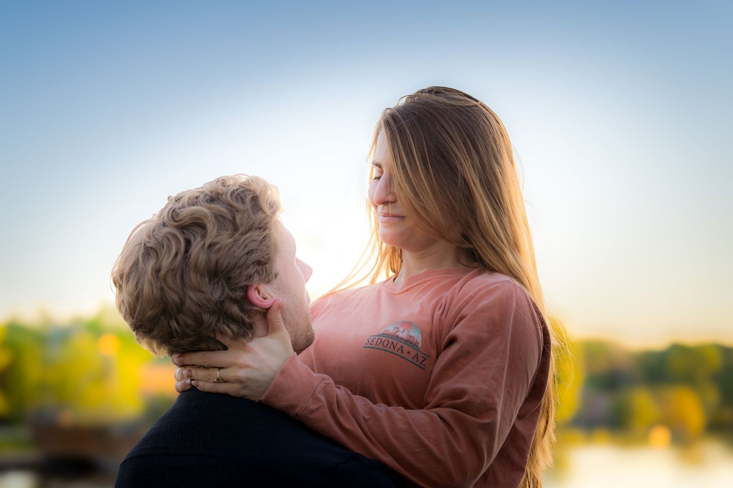 Woman with closed eyes being held by a man near a lake at sunset in a tender embrace.