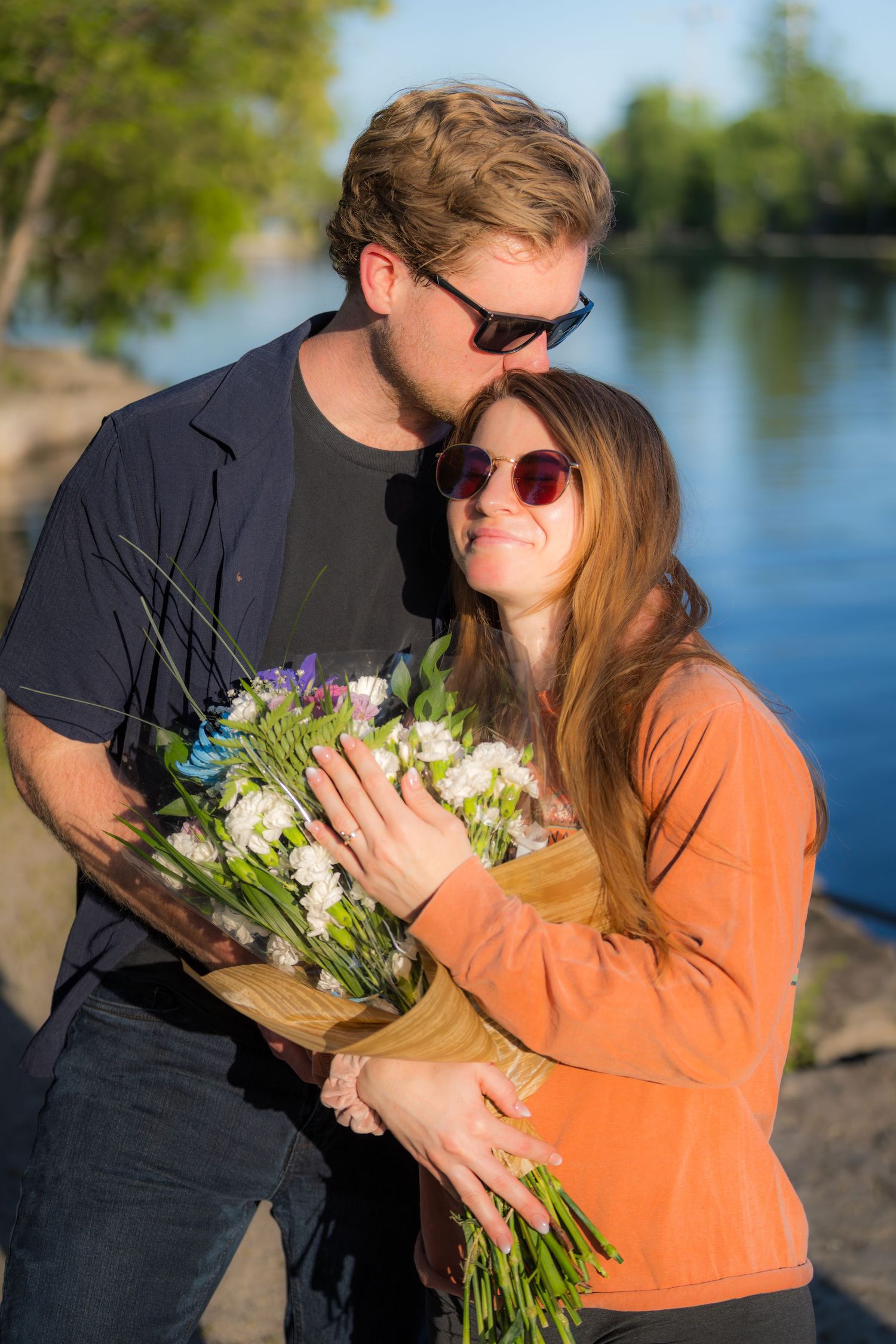 Happy couple embracing by a river, woman holding a bouquet while man kisses her cheek.