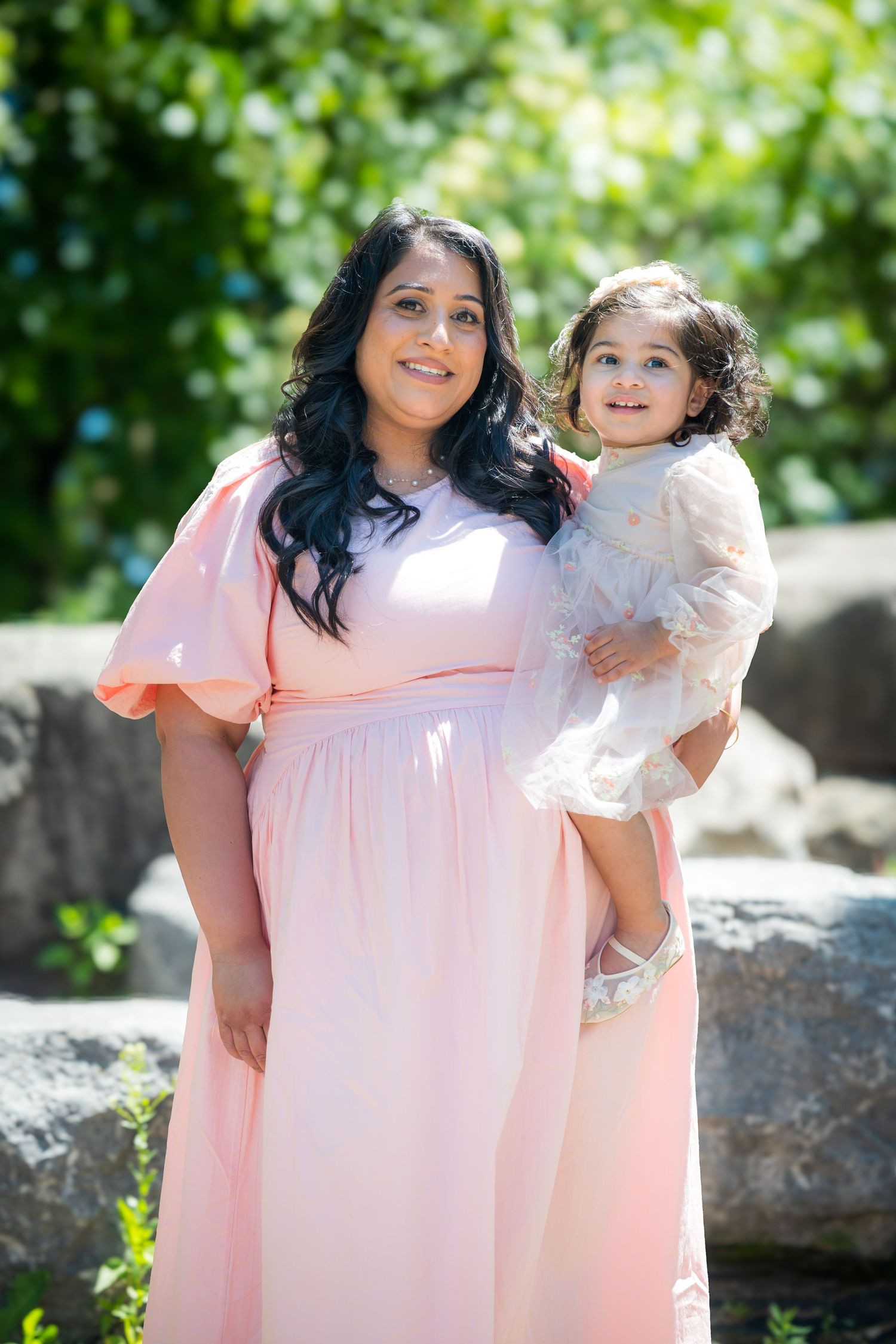 A mother in a pink dress holds her toddler daughter outdoors near rocks and green trees.
