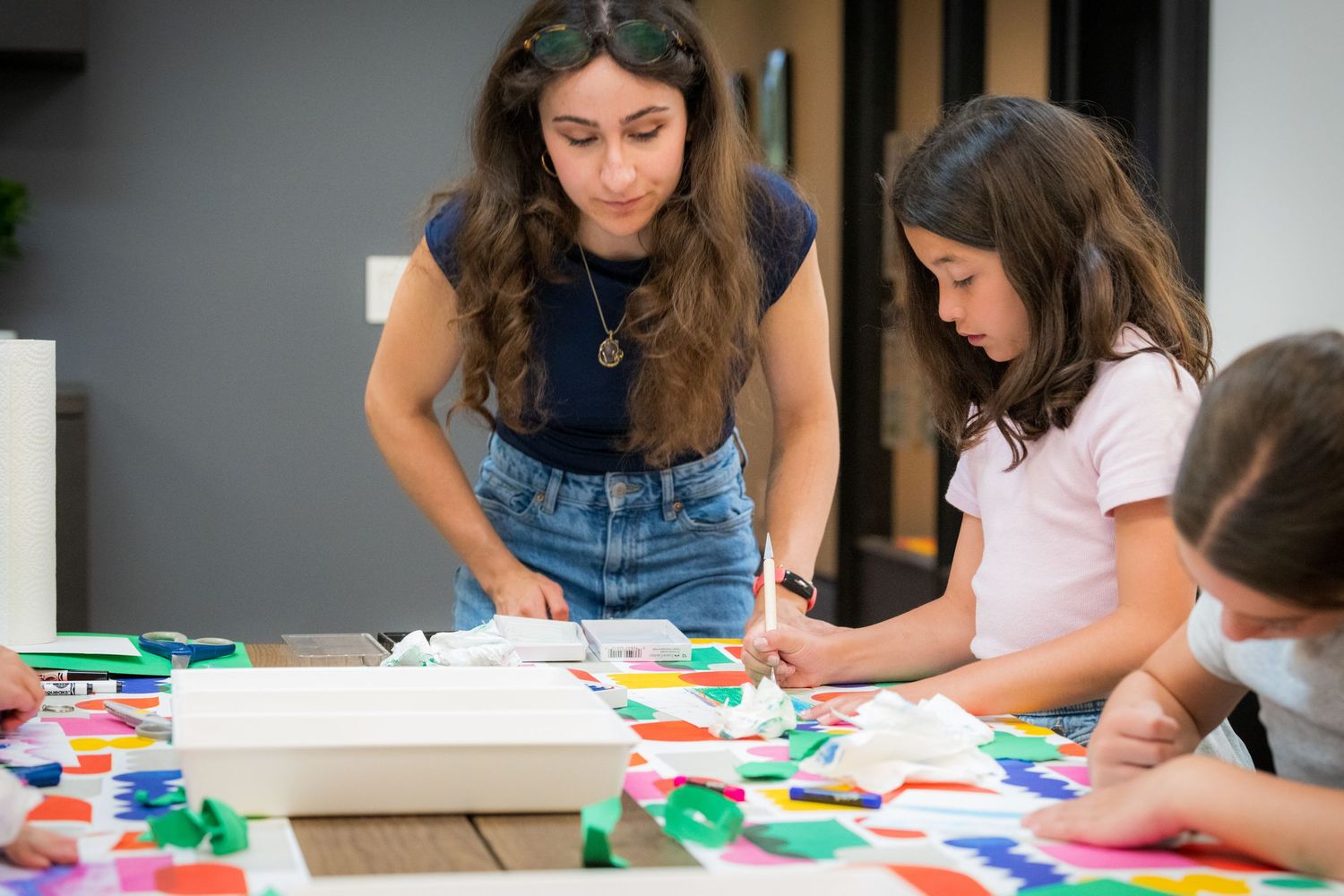 A woman assists two children with a colorful paper collage craft project at a table.