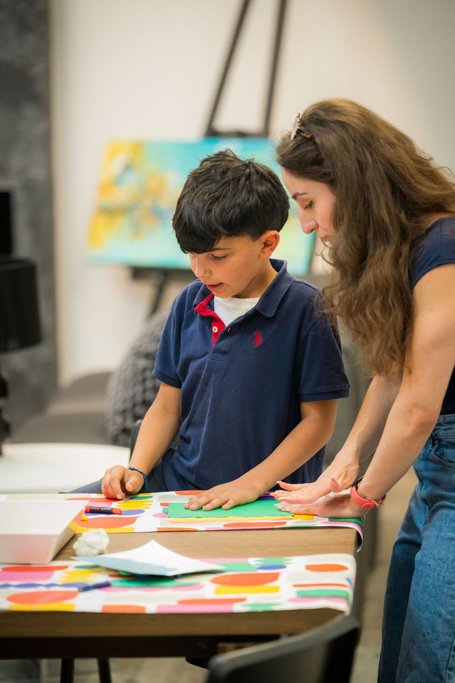A woman guides a young boy in a colorful paper craft art project at a table in an art gallery.