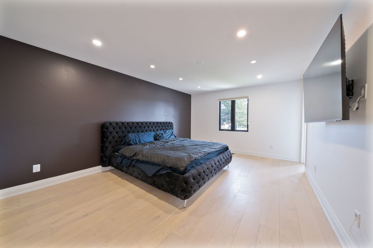 Modern bedroom with black tufted bed, dark accent wall, light hardwood floors and recessed lighting.