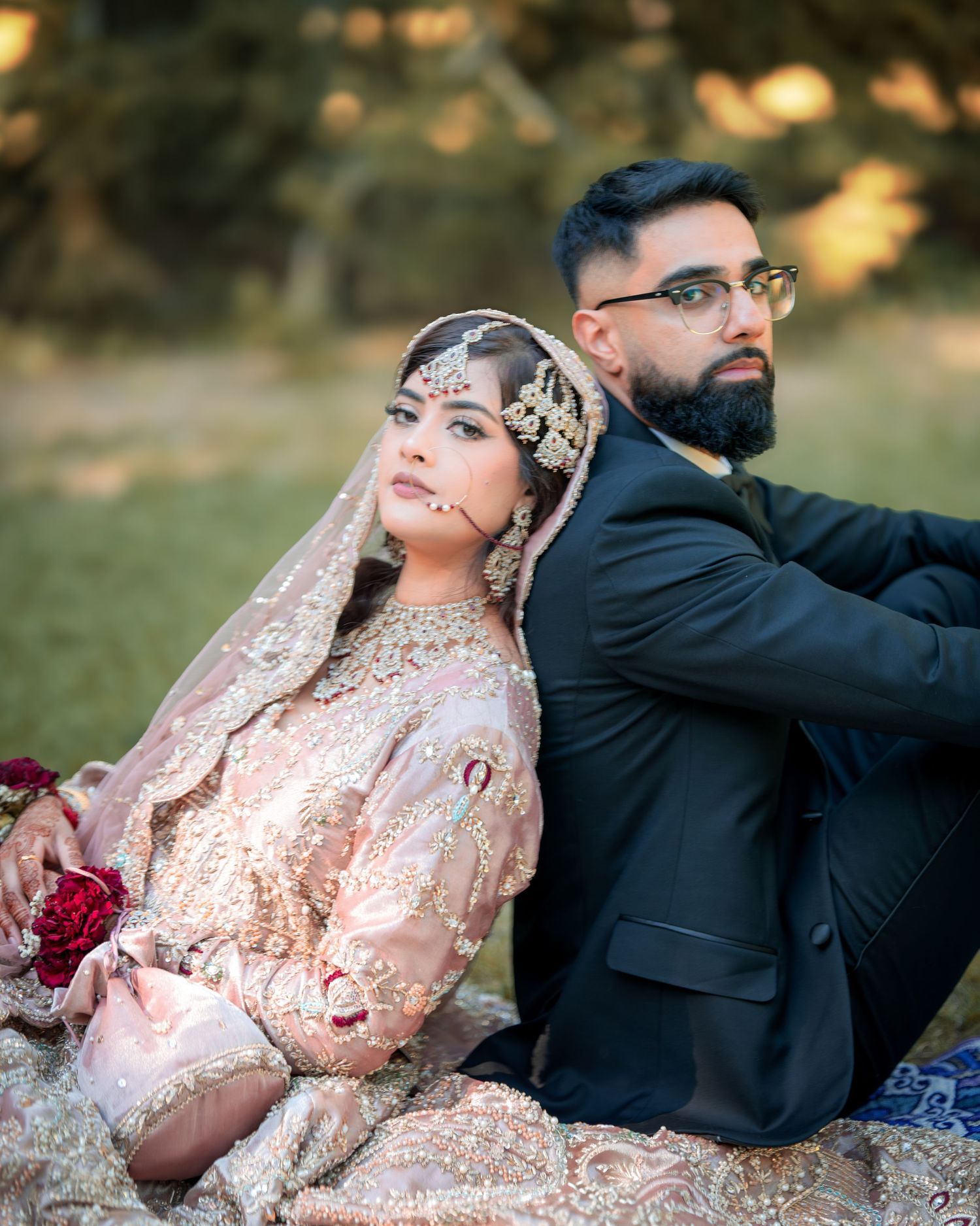 Wedding couple in traditional South Asian attire pose on grass during golden hour photoshoot.