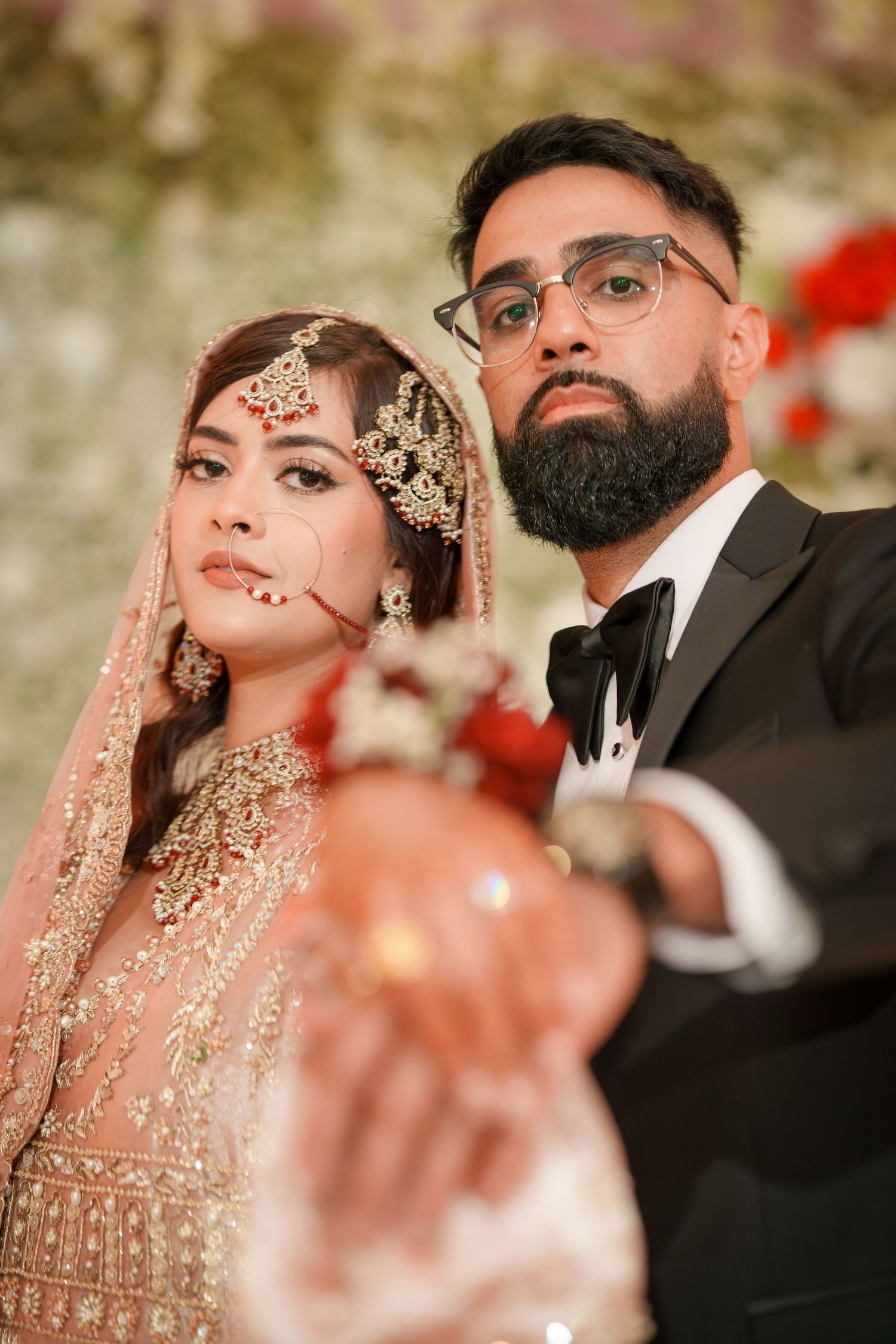 Couple in ornate South Asian wedding attire pose against floral backdrop.