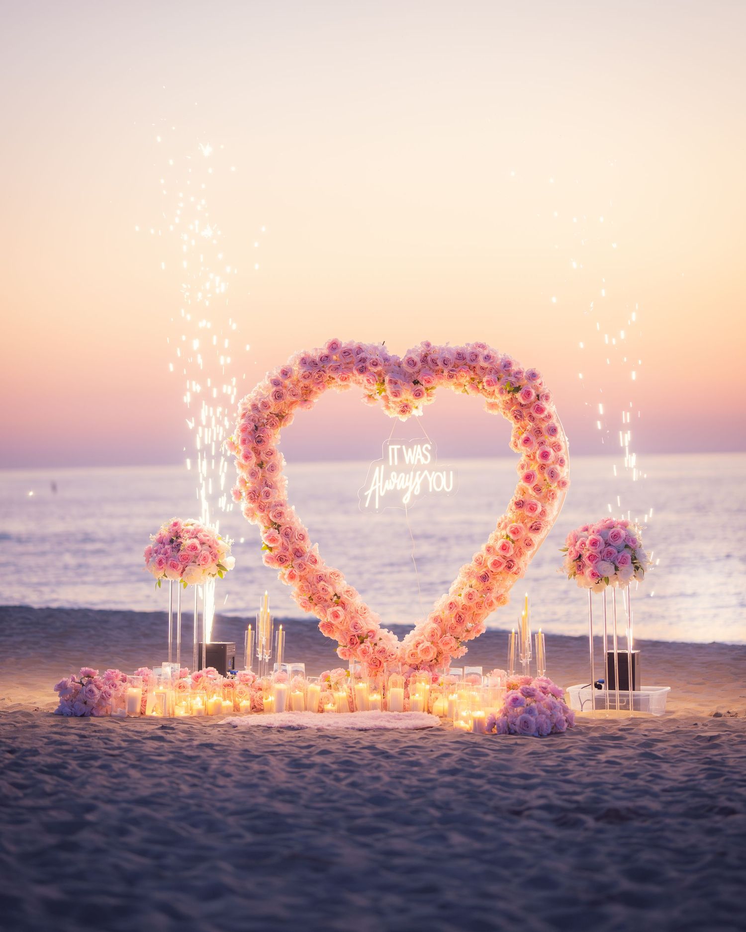 Beach proposal setup with heart-shaped flower arch and sparklers at dusk.