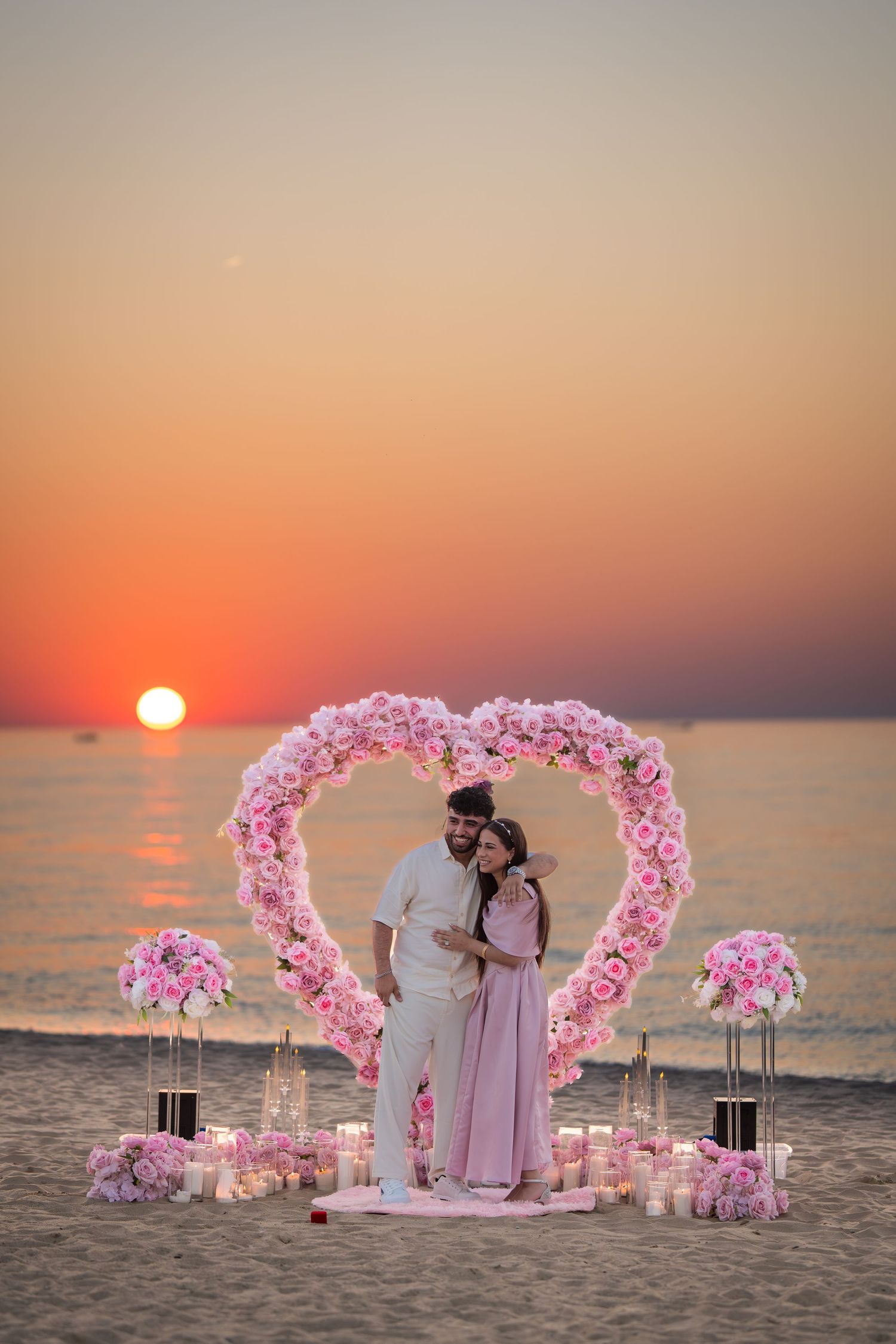 A dreamy beachside proposal scene with pink heart-shaped flower arch and glowing sunset over water.