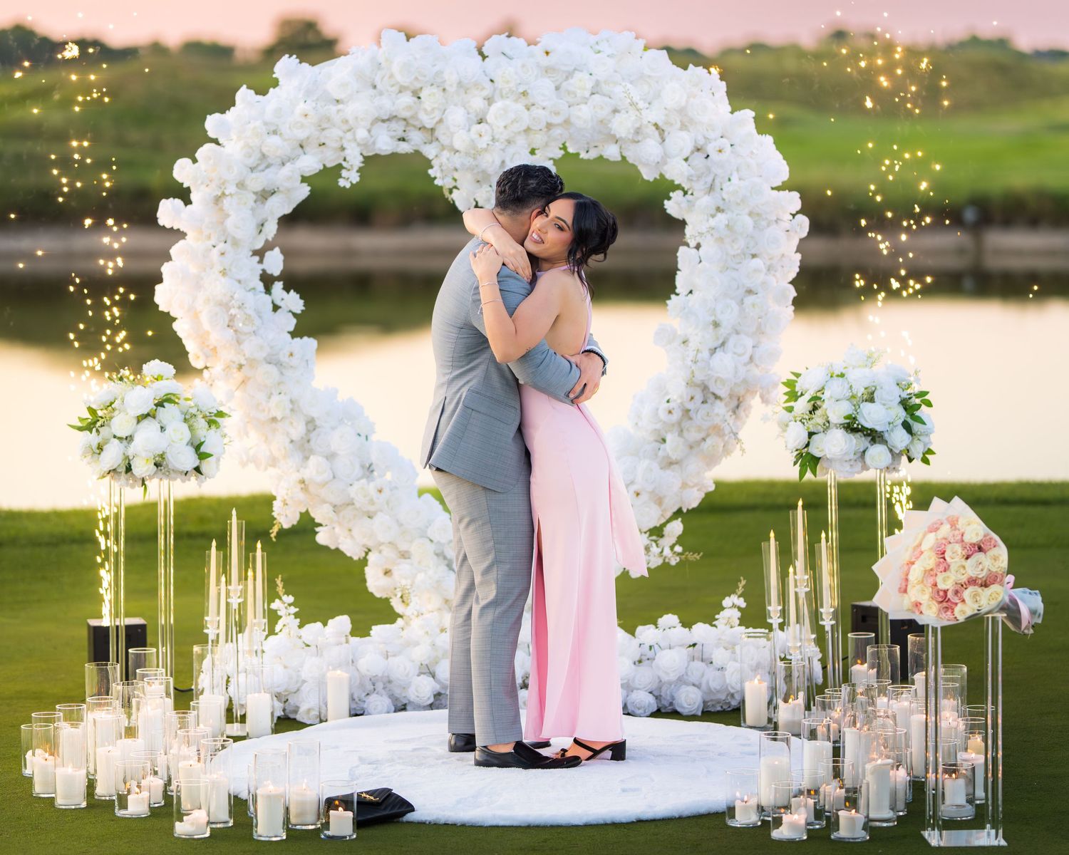 Beautiful lakeside proposal setting with white flower arch and scattered candles at dusk.
