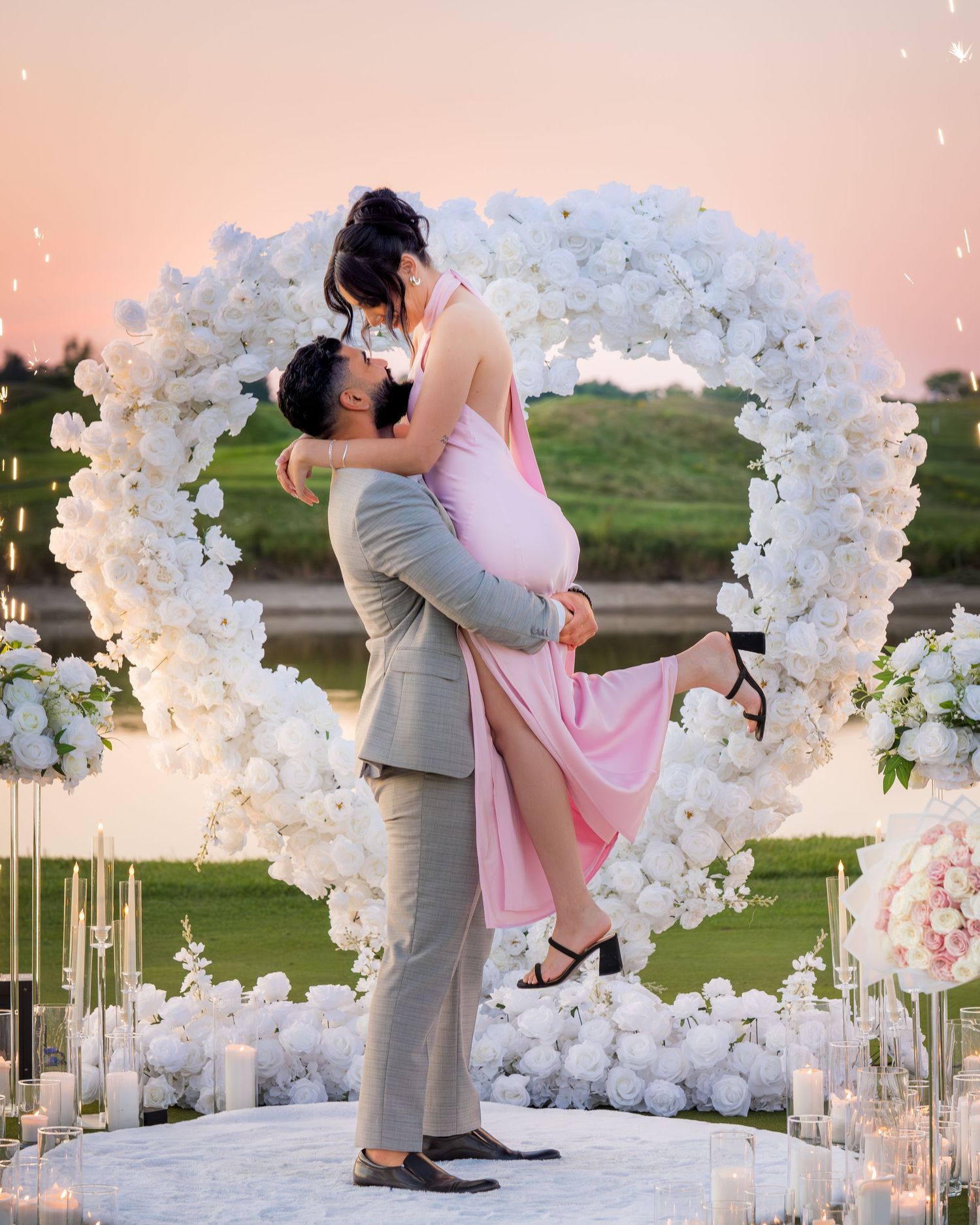 Romantic wedding pose captured beneath a white floral arch at golden hour.