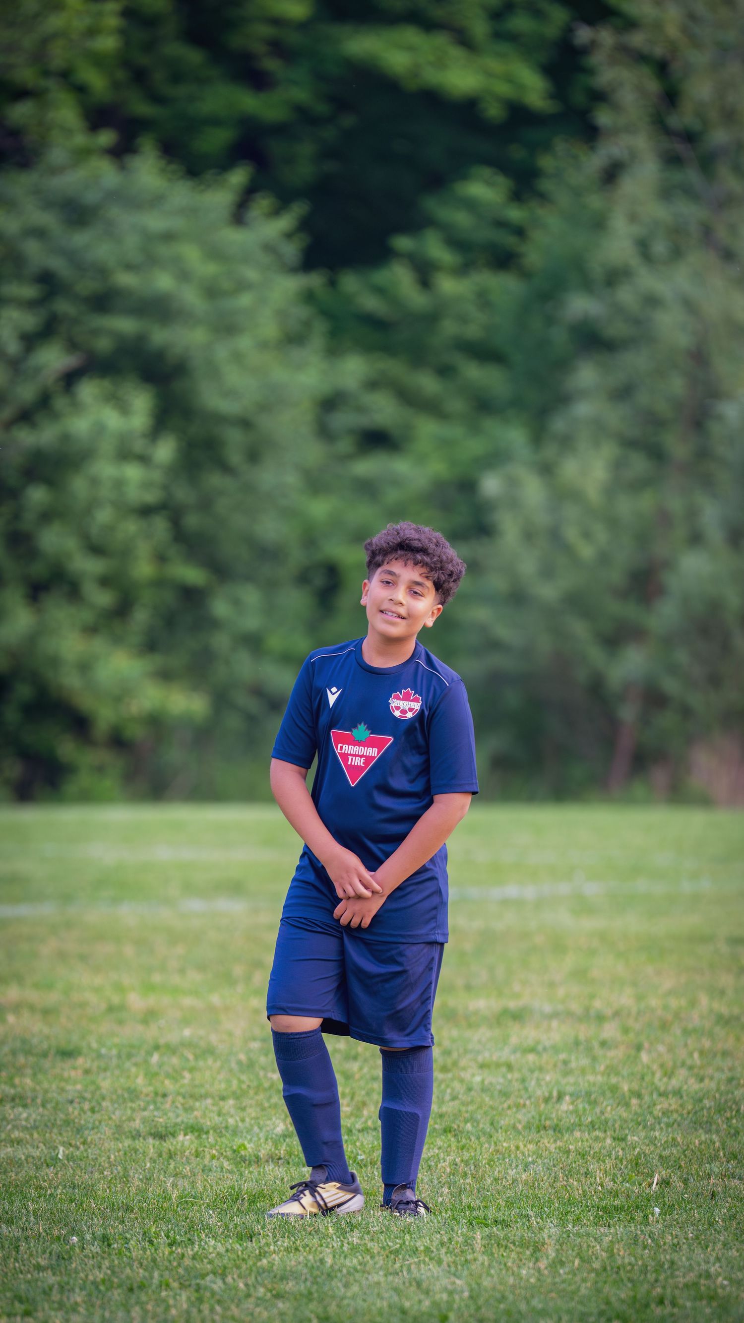 A youth soccer player in a navy blue uniform practices on a grassy field surrounded by trees.