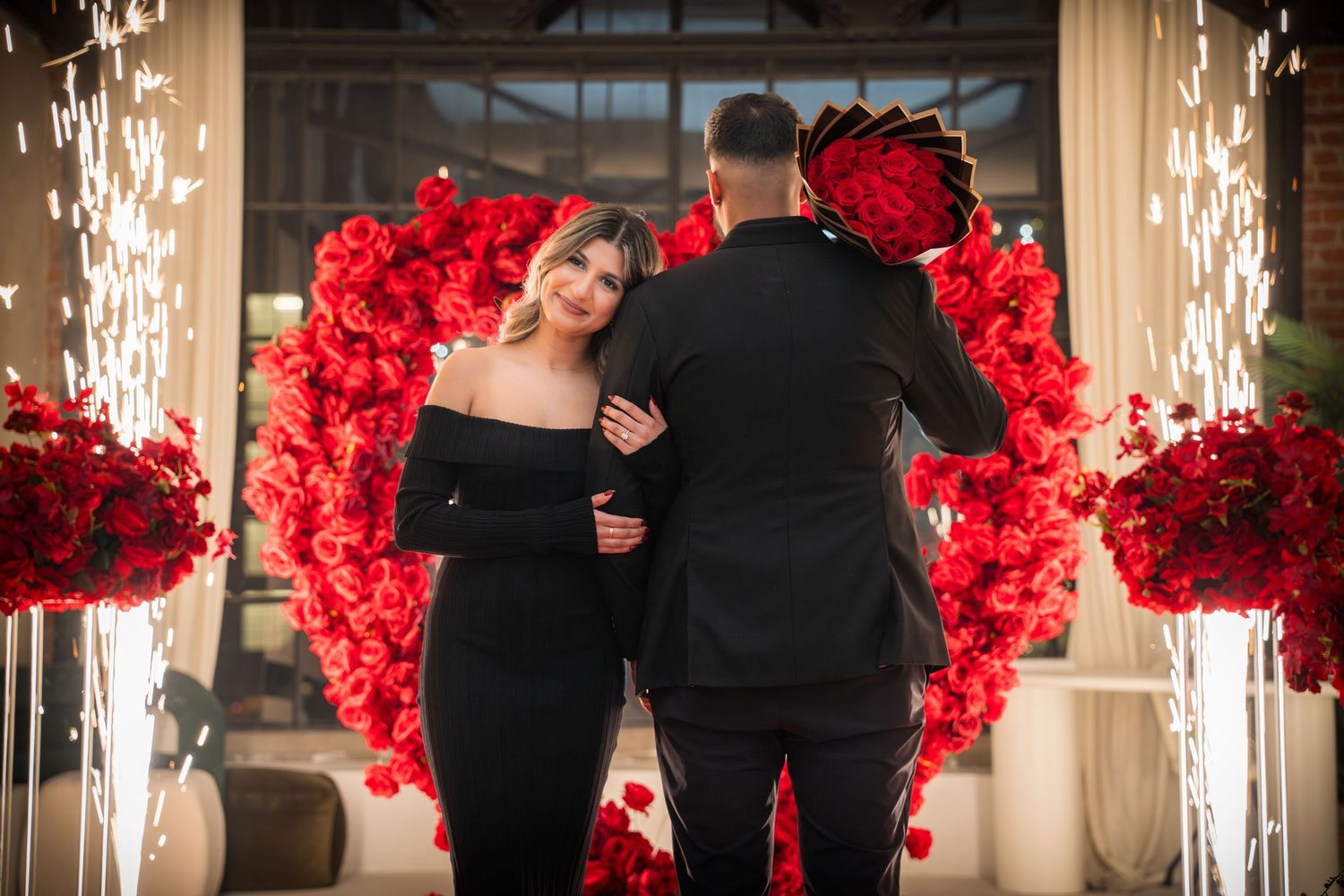 Couple stands in front of red rose heart-shaped arch with sparklers glowing in an elegant venue.