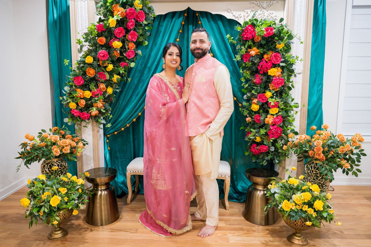 Couple in coordinating pink and cream traditional Indian outfits posing at colorful floral Lohri backdrop.