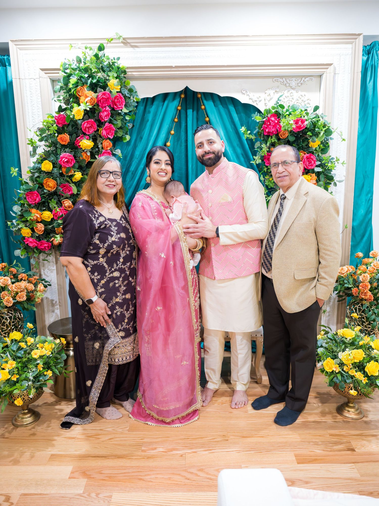 South Asian family of four in traditional dress poses with infant at floral decorated ceremony.