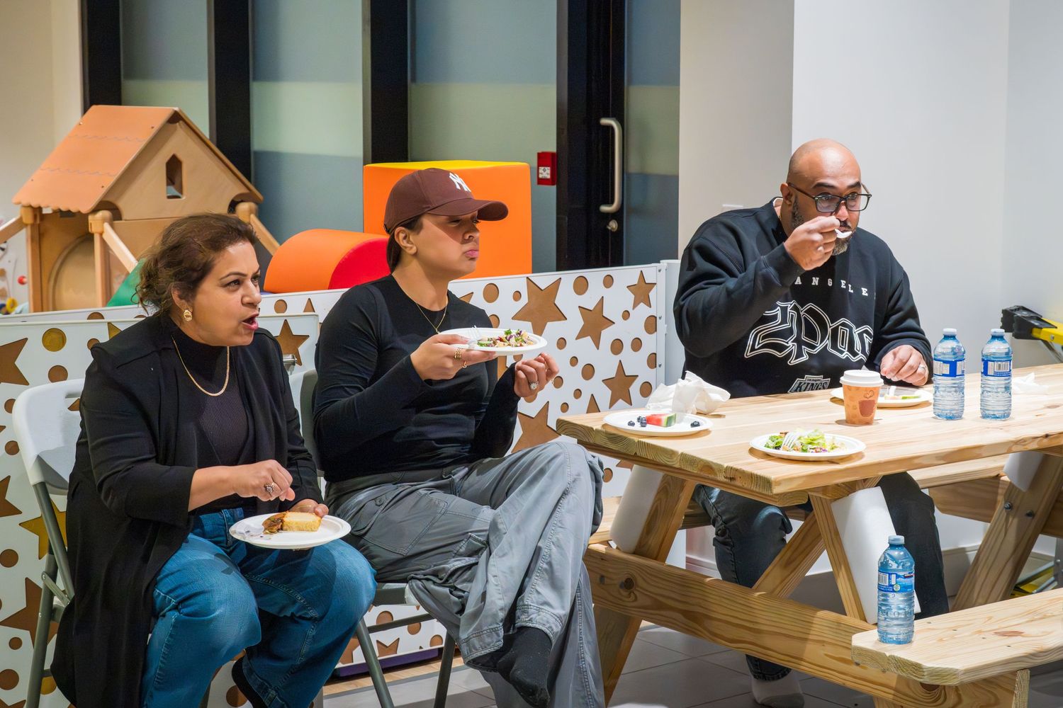 Three adults sit at a wooden picnic table eating food at an indoor party venue.