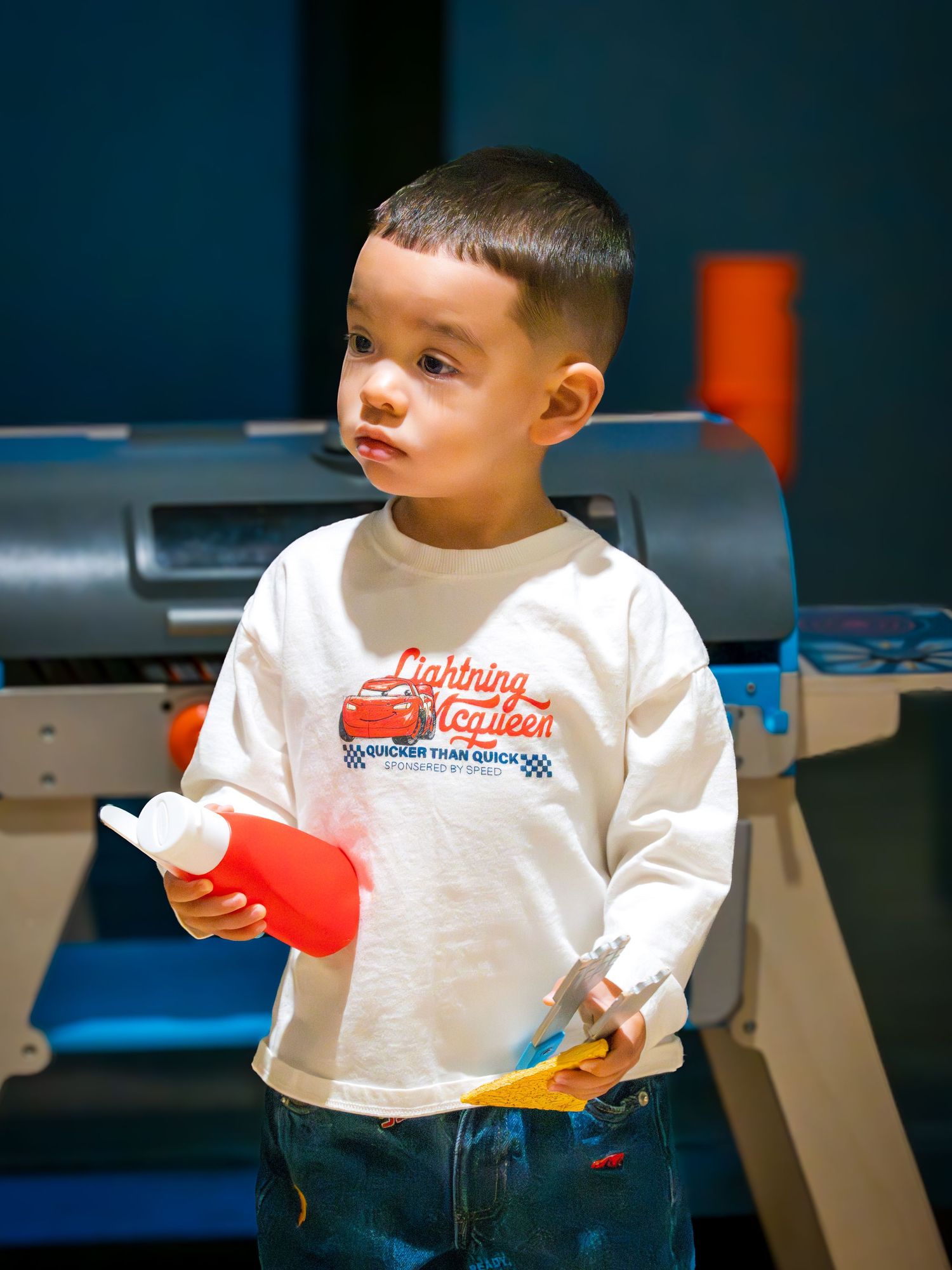 Toddler boy in a Lightning McQueen long-sleeve shirt holding toy tools at an indoor children's play area.