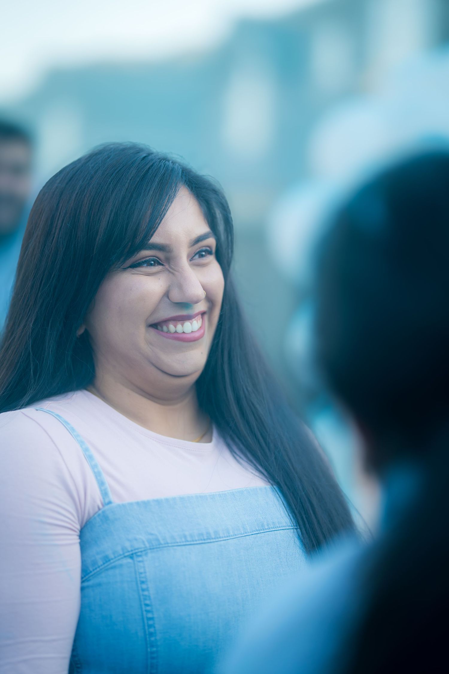 Smiling young woman in pink top and denim overalls at an outdoor gender reveal party.