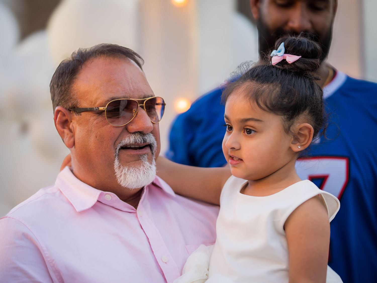 An older man with sunglasses holds a young girl in a white dress at an outdoor celebration.