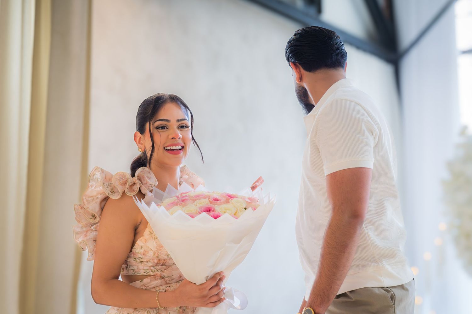 Joyful woman in a floral dress receiving a large bouquet of pink and white flowers from her partner indoors.