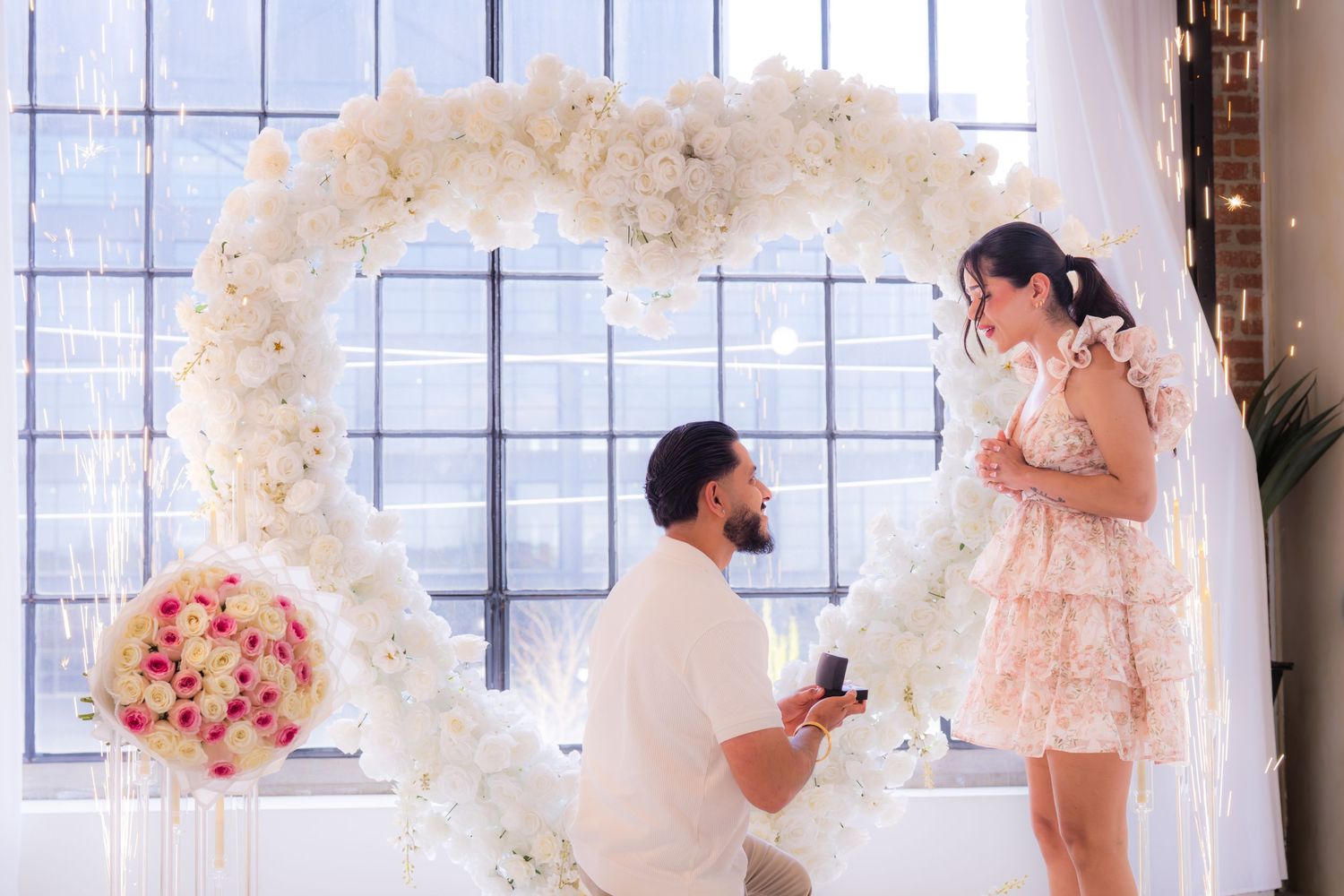Man on one knee proposing to his girlfriend under a white floral heart arch.